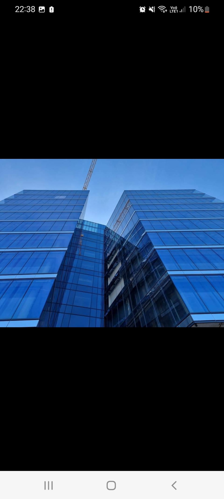 Niski kąt ujęcia na dwa nowoczesne, szklane biurowce w kolorze niebieskim, połączone przeszklonym łącznikiem, z widocznym żurawiem budowlanym w tle.