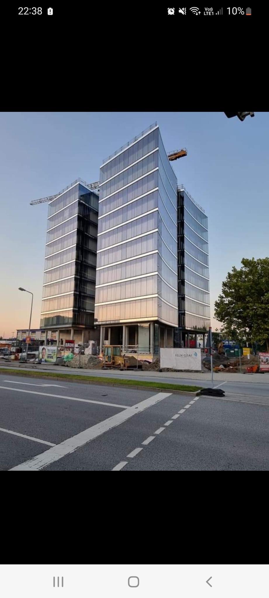 Dwa nowoczesne, szklane biurowce w trakcie budowy, widoczne z poziomu ulicy, z żółtą koparką i materiałami budowlanymi na placu budowy.