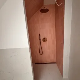 Nowoczesna kabina prysznicowa z różowymi, podłużnymi płytkami ułożonymi pionowo, mosiężna armatura i odpływ liniowy, surowy kabel wystający z sufitu.