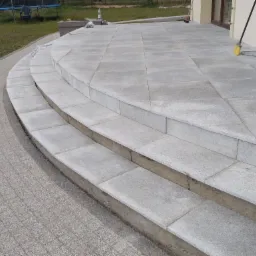 Granitowe schody wejściowe do domu z kostki, prowadzące na taras wyłożony płytami, widoczne z boku, z miotłą opartą o ścianę budynku.