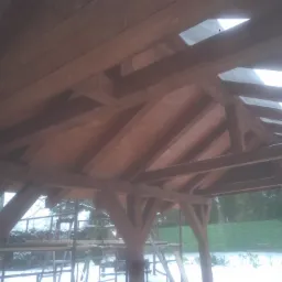 Drewniana konstrukcja dachu altany w trakcie budowy, widok od wewnątrz, z widocznymi elementami rusztowania na zewnątrz w zimowej scenerii.