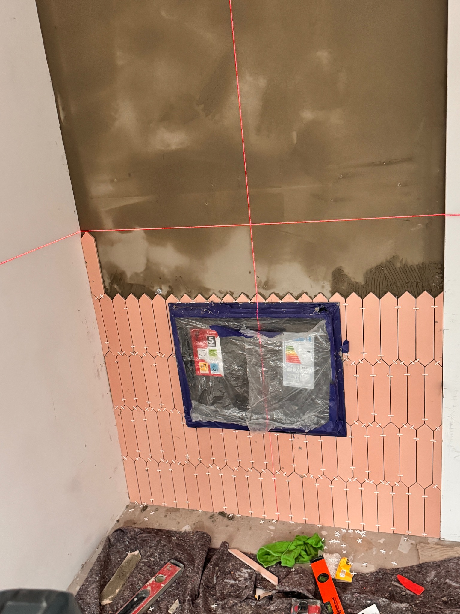 Układanie różowych płytek w kształcie strzałek na ścianie pod skosem, widoczne krzyżyki dystansowe, poziomica i laserowy wskaźnik pionu, okno zabezpieczone folią.