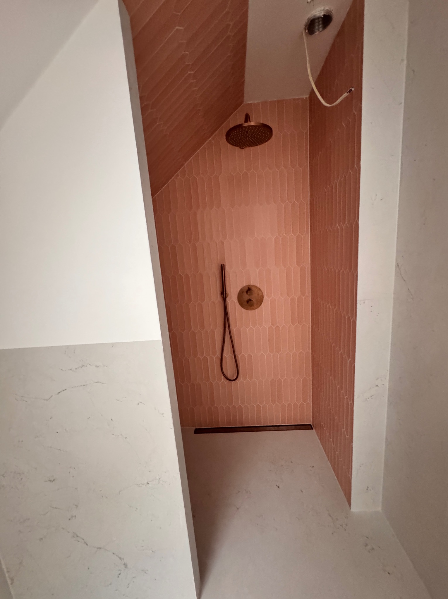 Nowoczesna kabina prysznicowa z różowymi, podłużnymi płytkami ułożonymi pionowo, mosiężna armatura i odpływ liniowy, surowy kabel wystający z sufitu.