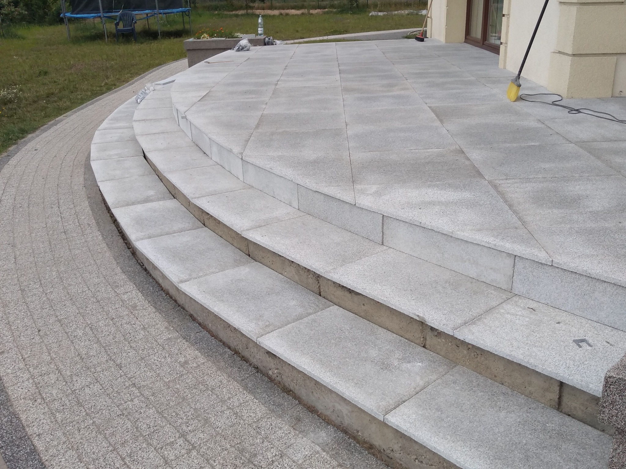 Granitowe schody wejściowe do domu z kostki, prowadzące na taras wyłożony płytami, widoczne z boku, z miotłą opartą o ścianę budynku.