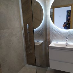 BudWork - Nowoczesna łazienka z kabiną prysznicową typu walk-in, umywalką z szafką w kolorze białym i okrągłym lustrem podświetlanym LED, odbicie fotografa w lustrze.