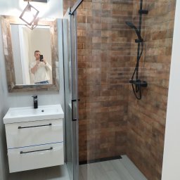 BudWork - Wykończona łazienka w Warszawie z kabiną prysznicową typu walk-in z czarnymi elementami, umywalką z szafką w bieli oraz lustrem z drewnianą ramą, w której odbija się osoba robiąca zdjęcie.