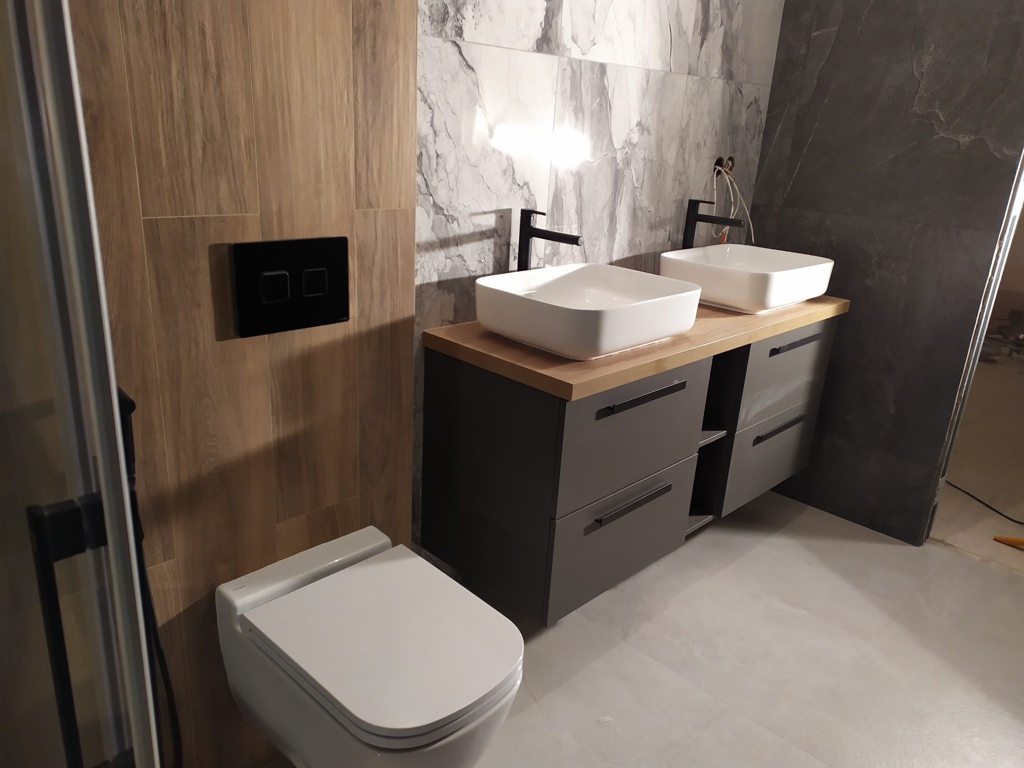 Nowoczesna łazienka z dwoma białymi umywalkami nablatowymi na szarej szafce z drewnianym blatem, czarne baterie, wisząca toaleta i płytki imitujące drewno oraz marmur.