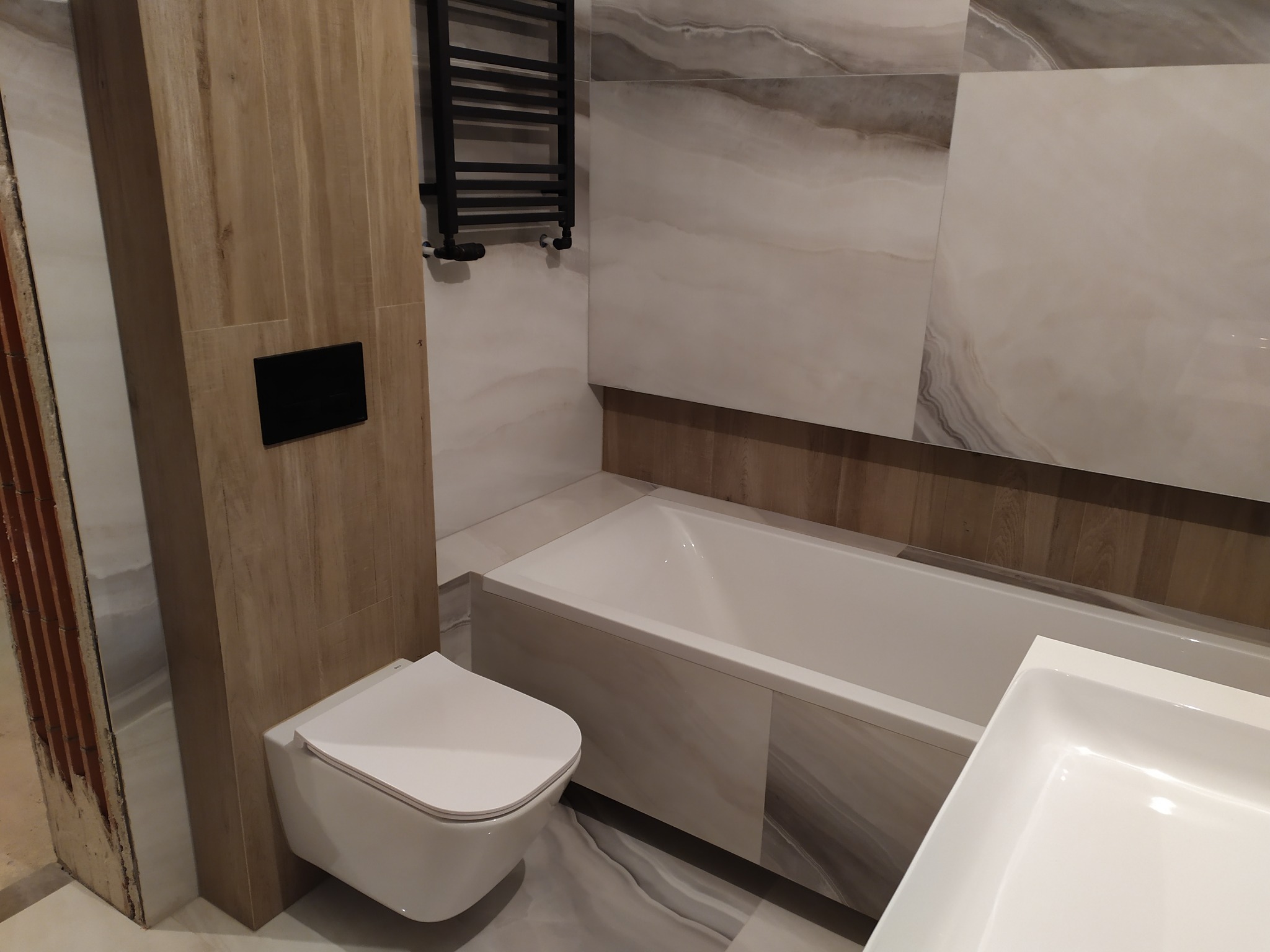 Nowoczesna łazienka po remoncie z białą armaturą, drewnopodobnymi płytkami i czarnym grzejnikiem drabinkowym.