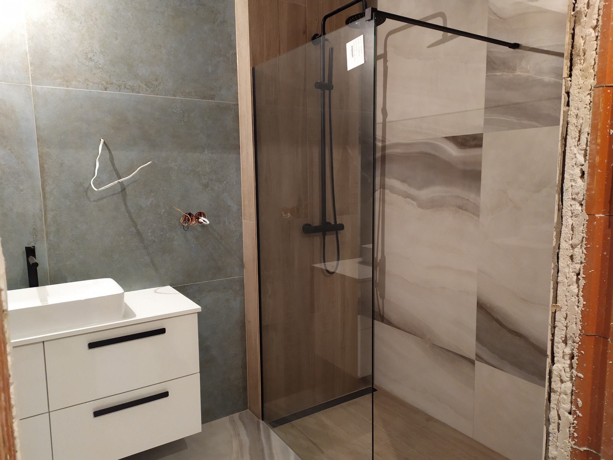 Nowoczesna łazienka w trakcie aranżacji: biała szafka podumywalkowa, szara ściana z wystającymi przewodami, kabina prysznicowa z czarną armaturą i płytkami imitującymi marmur, widoczne...