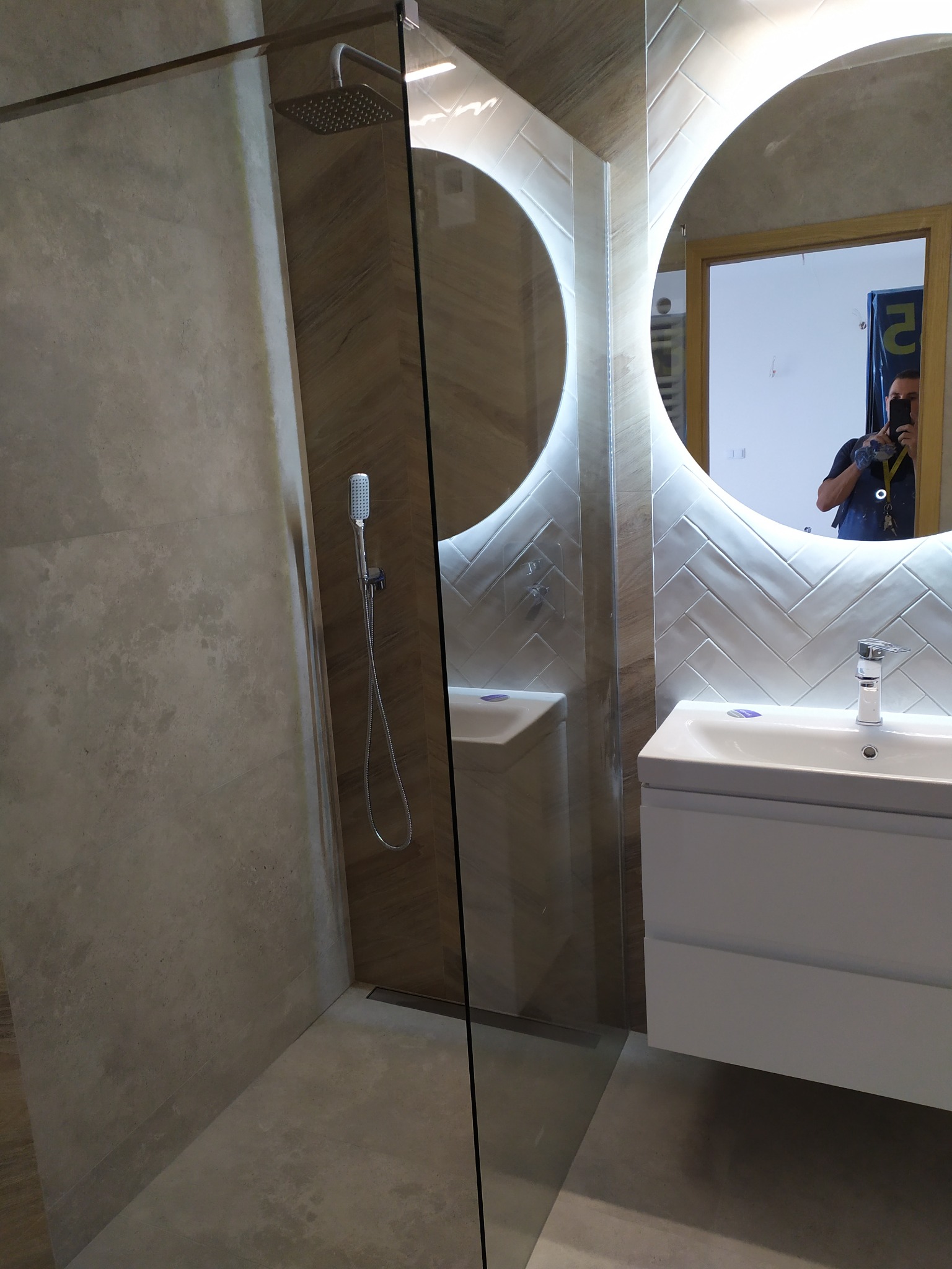 Nowoczesna łazienka z kabiną prysznicową typu walk-in, umywalką z szafką w kolorze białym i okrągłym lustrem podświetlanym LED, odbicie fotografa w lustrze.