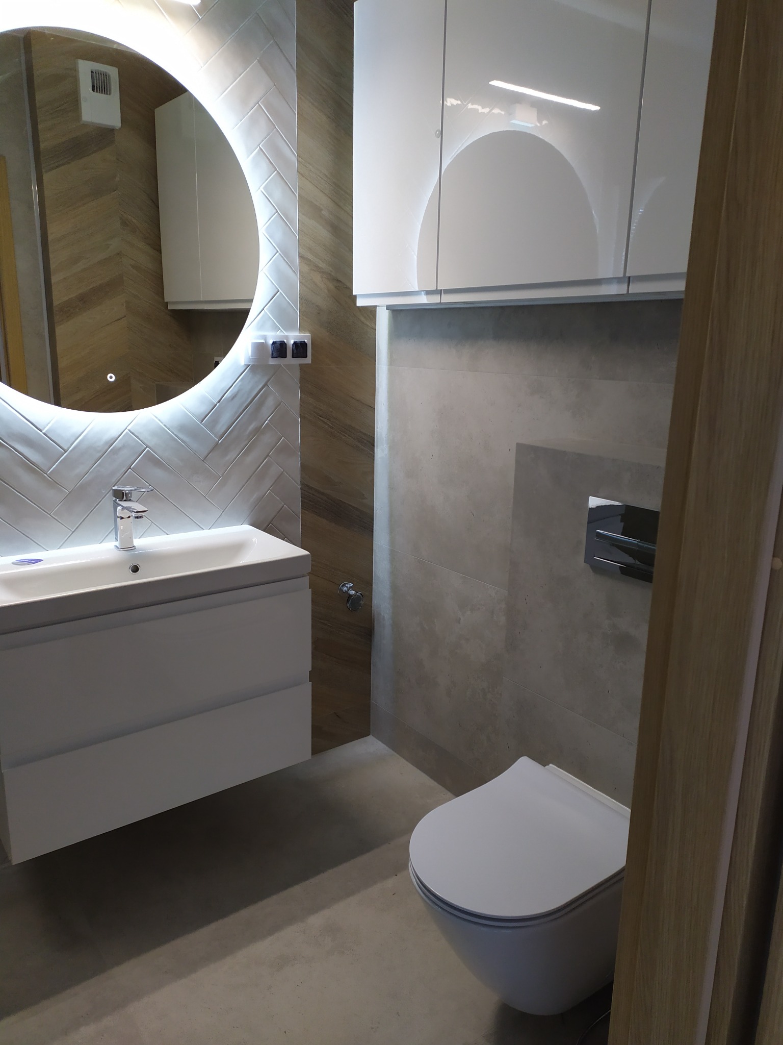 Nowoczesna łazienka z podświetlanym okrągłym lustrem, białą umywalką z szafką, wiszącą toaletą i szarymi płytkami na ścianach.
