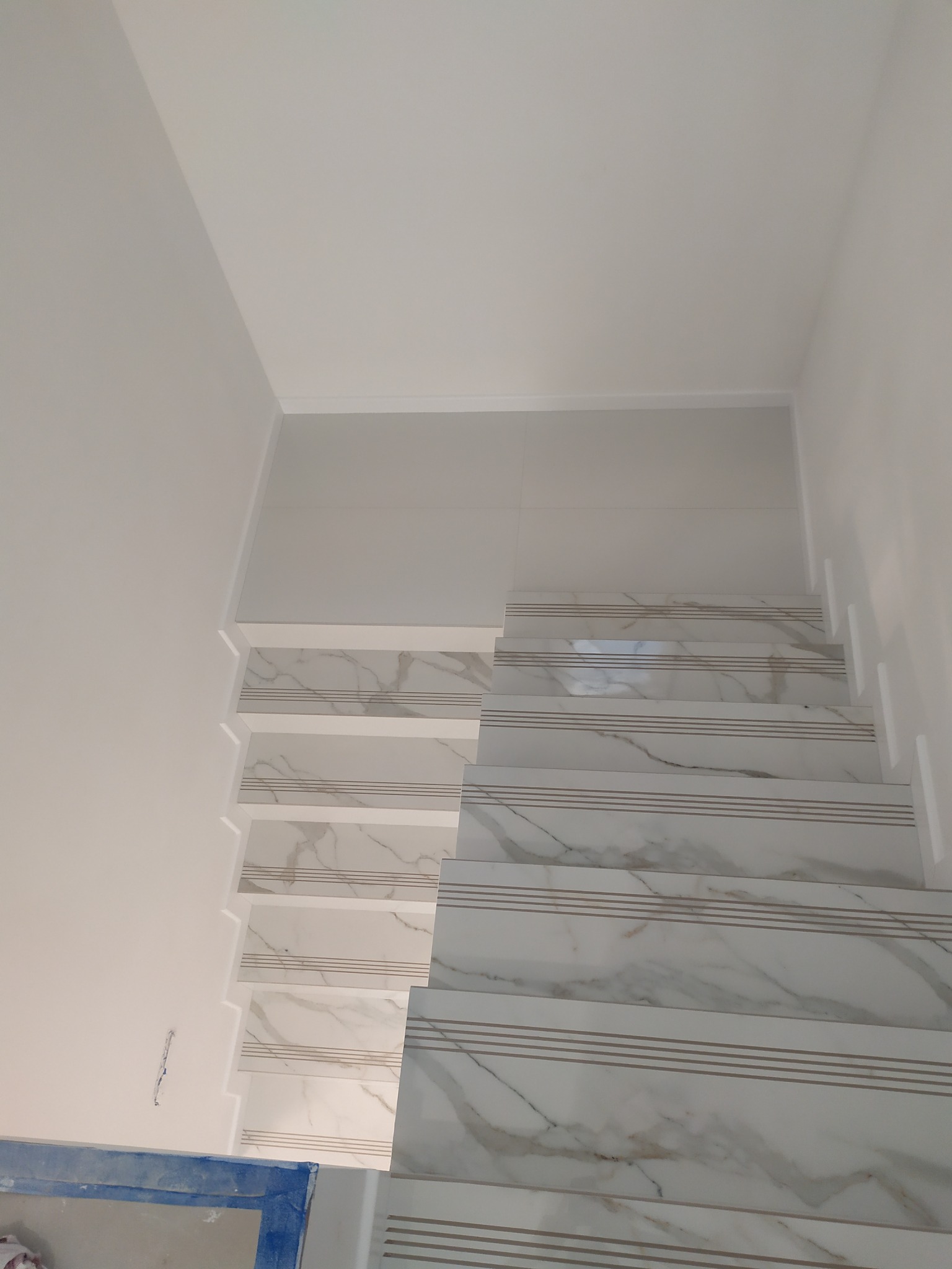 Wykończone schody wewnętrzne z białymi płytkami imitującymi marmur, z trzema poziomymi paskami, widok z dołu w górę, fragment niebieskiego pojemnika w lewym dolnym rogu.