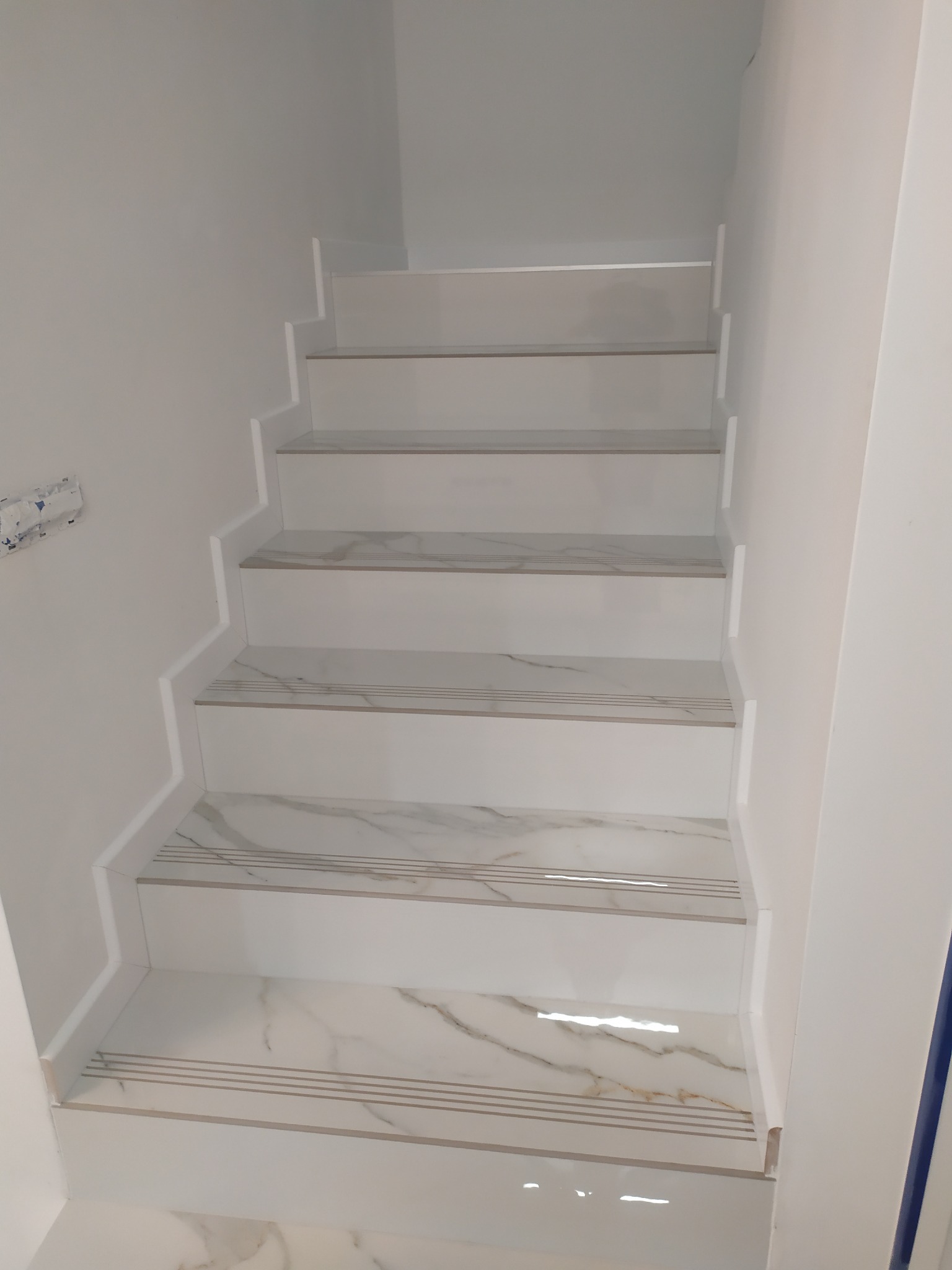 Wykończone schody wewnątrz budynku, wyłożone białymi płytkami z marmurkowym wzorem i trzema poziomymi paskami antypoślizgowymi na krawędziach stopni.