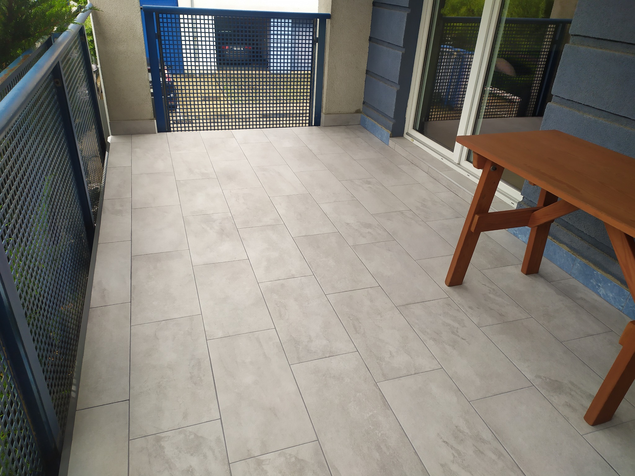 Wyłożony jasnoszarą płytką balkon z metalową barierką i drewnianym stolikiem.