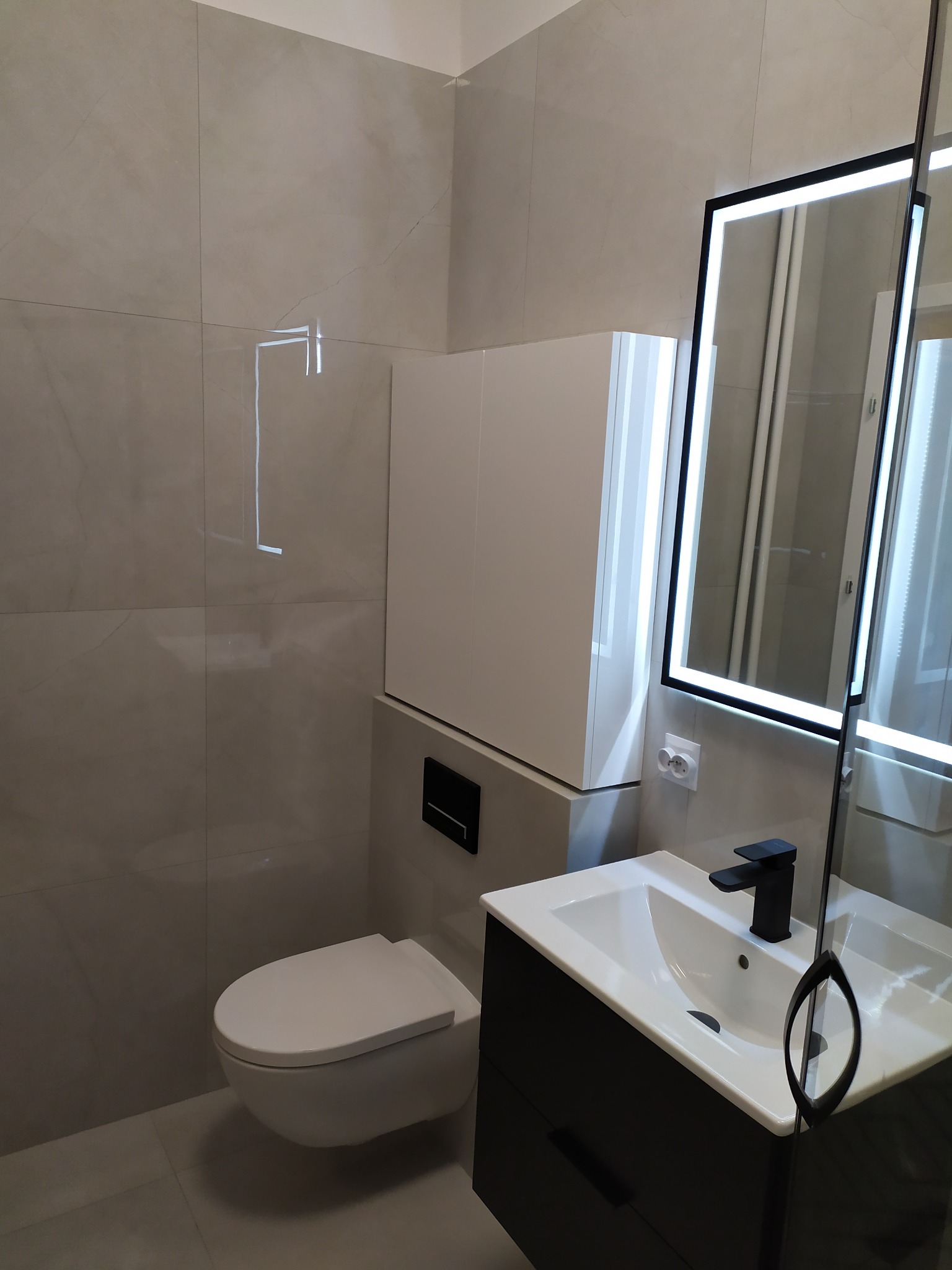 Nowoczesna łazienka z szarymi płytkami, podwieszaną toaletą i szafką, umywalką z czarną armaturą oraz lustrem z podświetleniem LED.