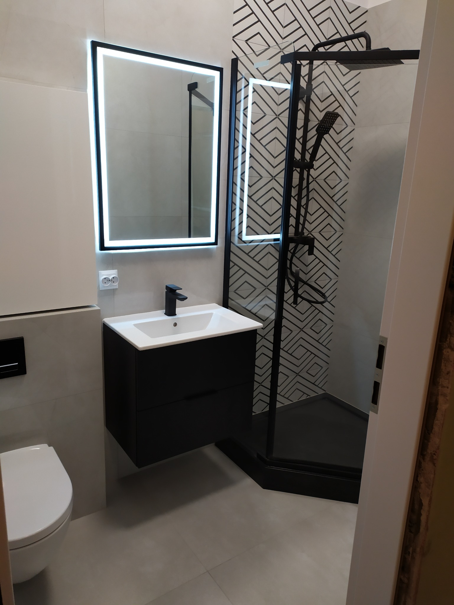 Nowoczesna łazienka z czarną armaturą: kabina prysznicowa z geometrycznym wzorem na ścianie, podświetlane lustro nad szafką umywalkową i wisząca toaleta.