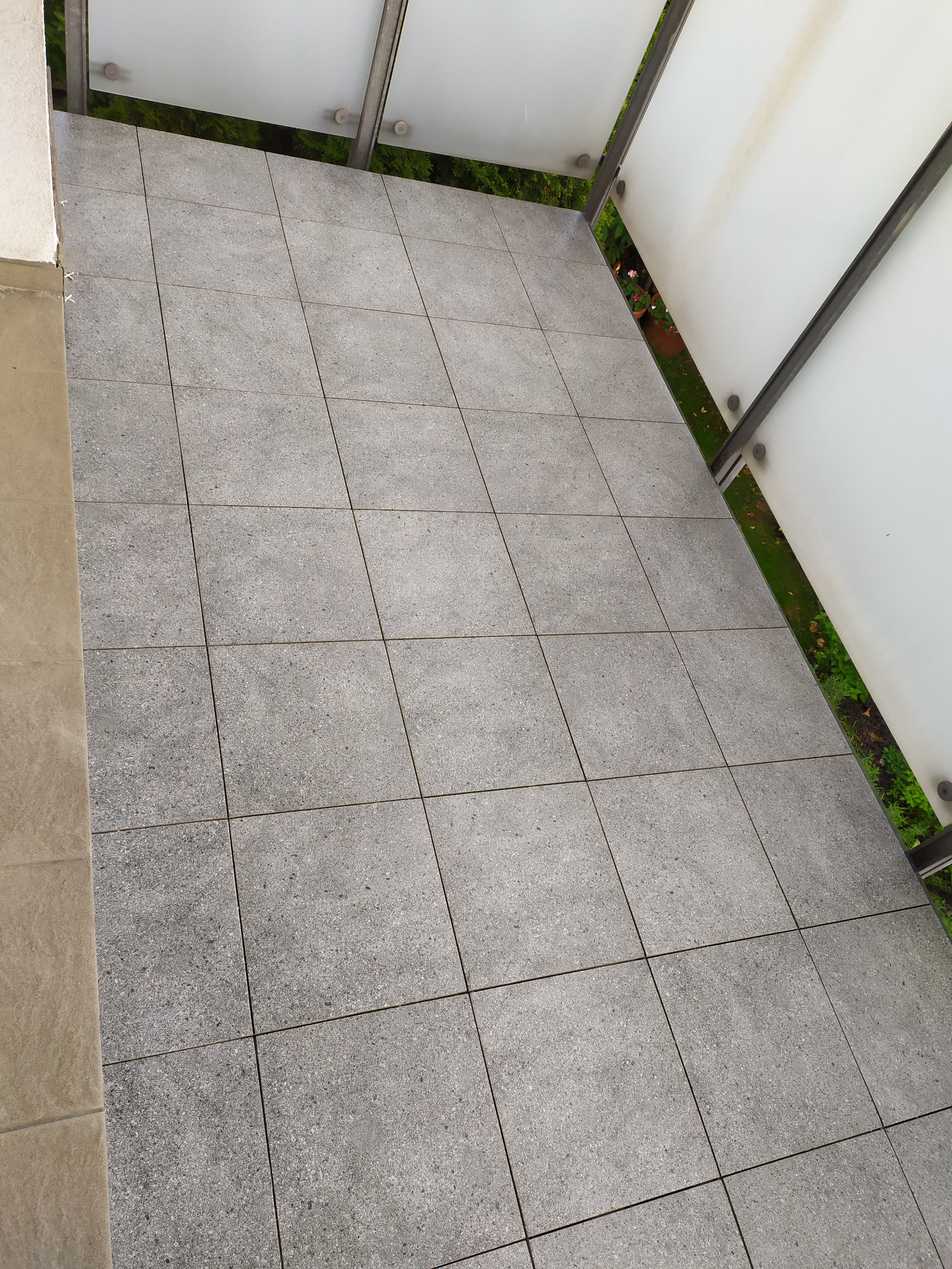 Szare płytki gresowe ułożone na balkonie, widoczna szklana balustrada i zieleń w tle.