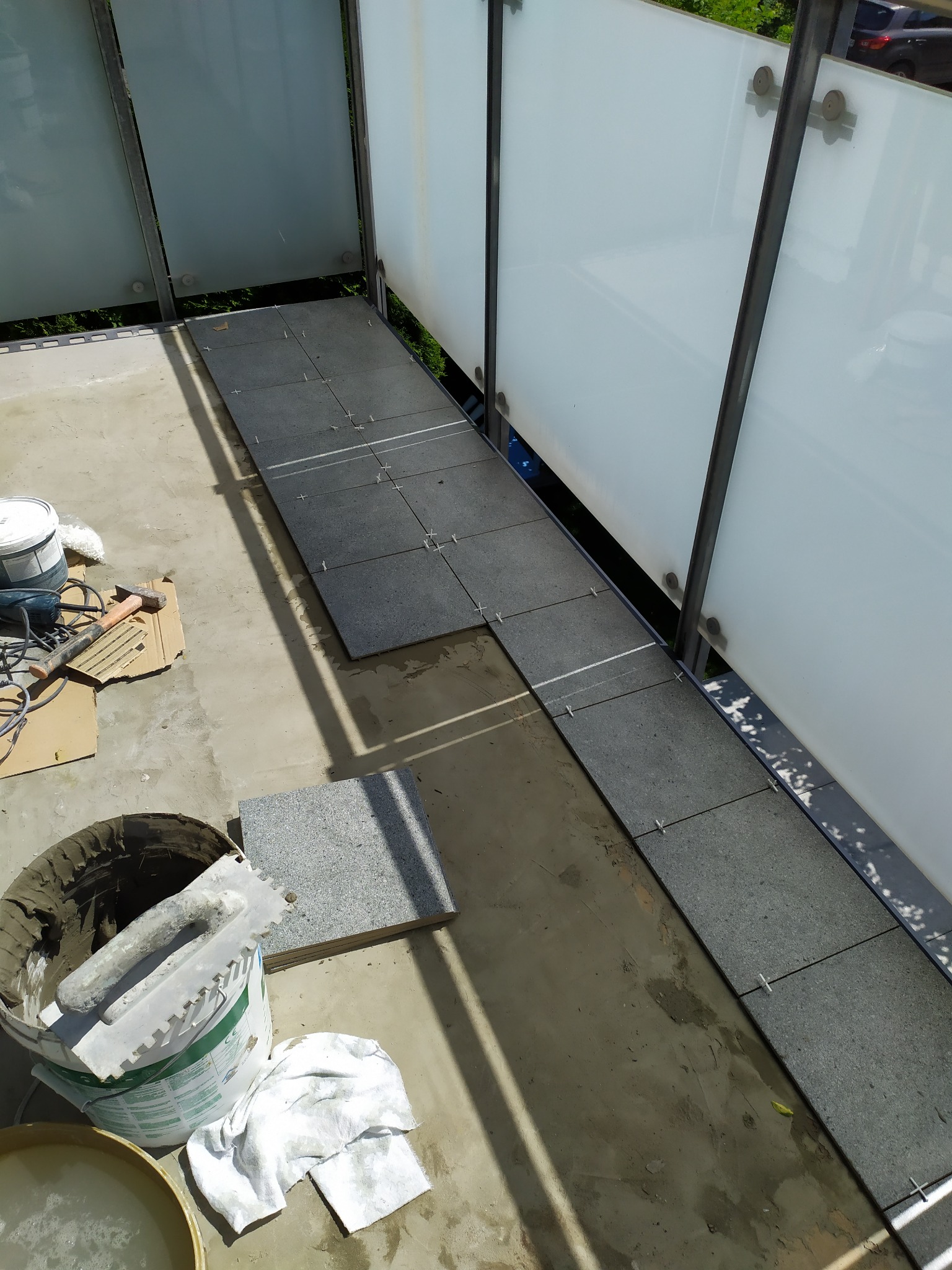 Montaż szarych płytek na balkonie z białymi, szklanymi balustradami, widoczne krzyżyki dystansowe, wiadro z zaprawą, kielnia i narzędzia.