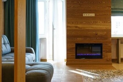 Salon z drewnianą ścianą, wbudowanym elektrycznym kominkiem z niebieskim płomieniem, szarą sofą i wzorzystą podłogą.