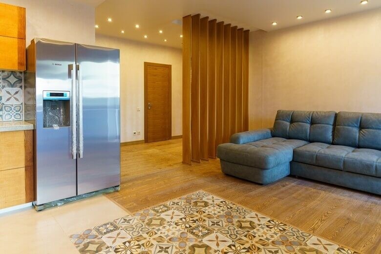 Nowoczesne wnętrze mieszkania z drewnianą ścianką działową, szarą sofą narożną, dwudrzwiową lodówką Bosch z kostkarką, podłogą z płytek w stylu patchwork i drewnianą podłogą.