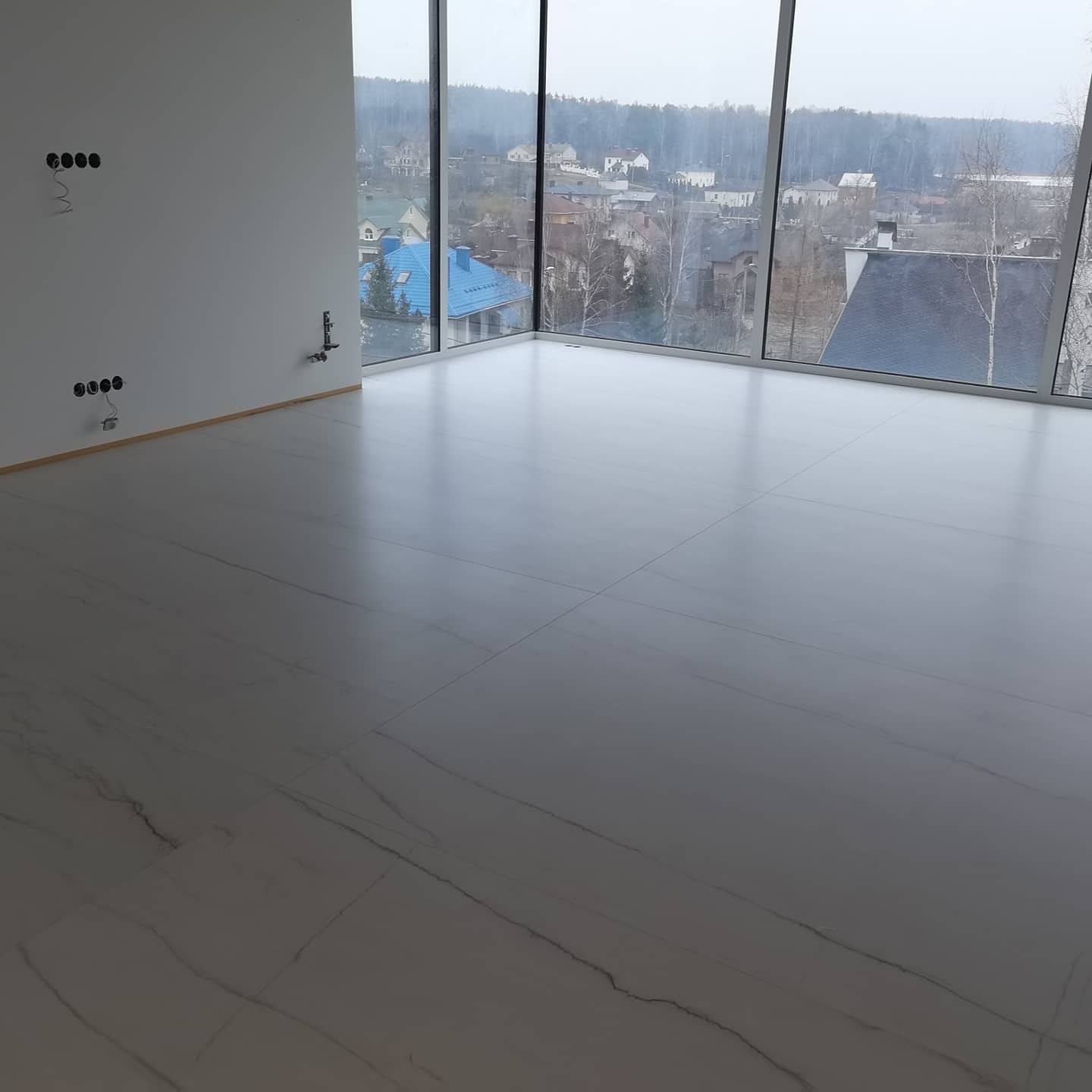 Jasna, marmurowa podłoga w nowym, przestronnym pokoju z dużymi oknami, widok na zabudowę miejską w tle i wystającymi przewodami elektrycznymi ze ściany.