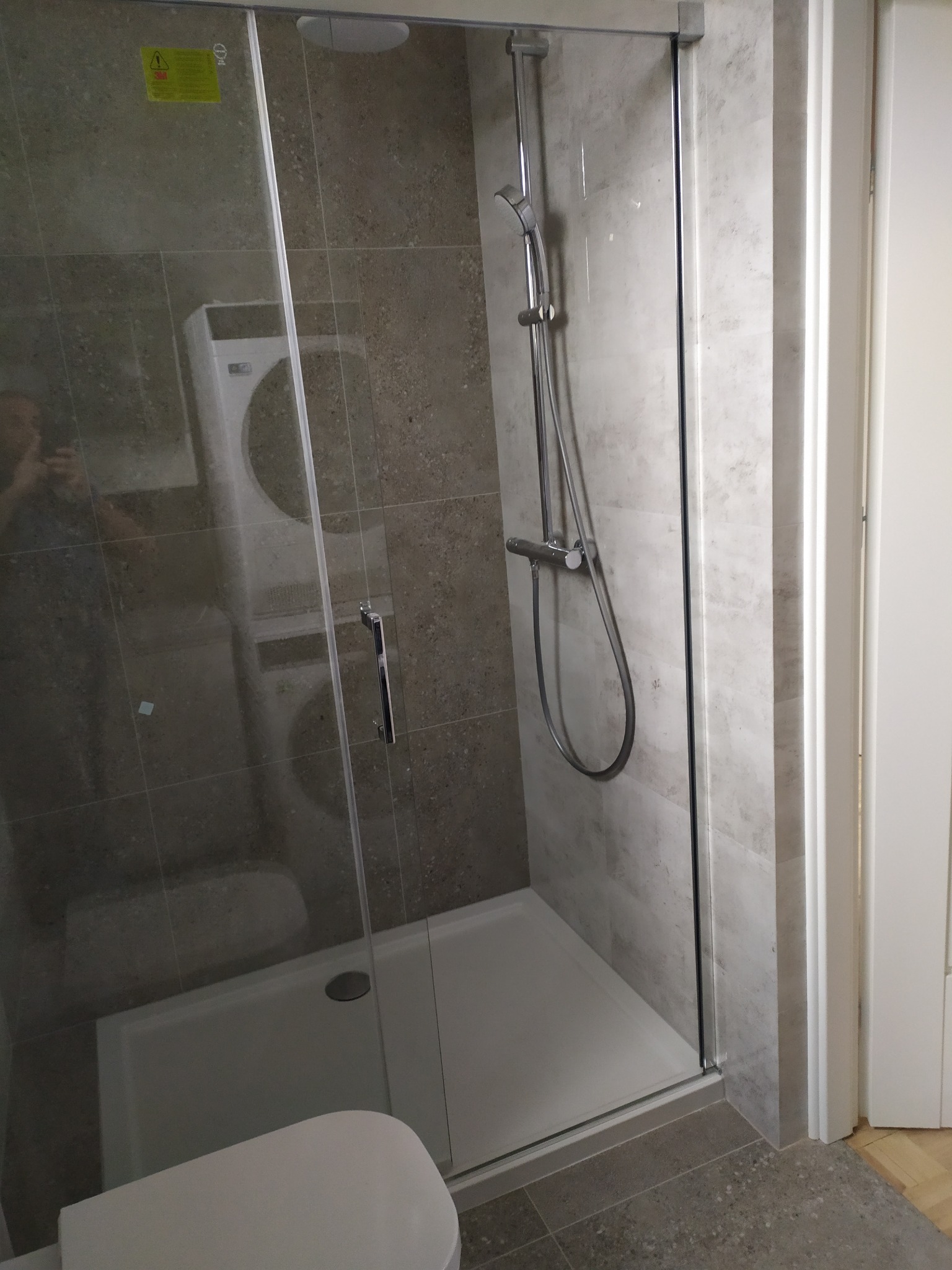 Nowoczesna kabina prysznicowa z szarymi płytkami imitującymi kamień, widoczna pralka w odbiciu szkła.