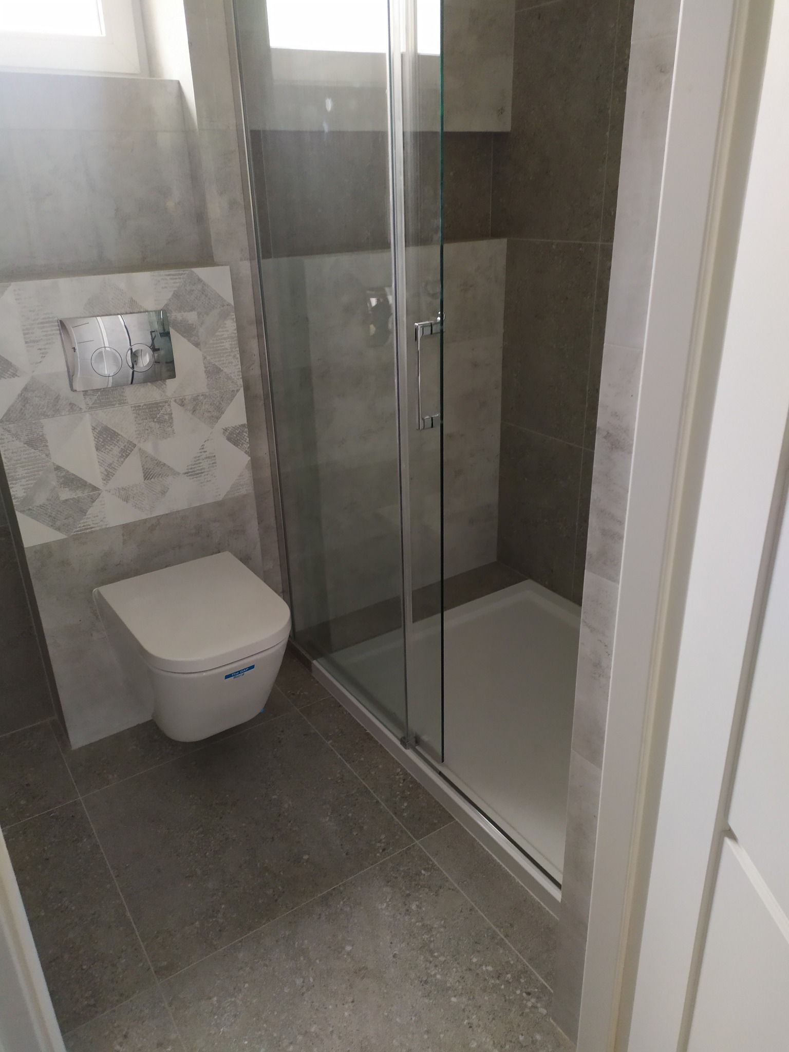 Nowoczesna łazienka z kabiną prysznicową za szklanymi drzwiami, podwieszaną toaletą z chromowanym przyciskiem spłukującym na ścianie pokrytej dekoracyjnymi płytkami w geometryczne wzory, oraz szarą...