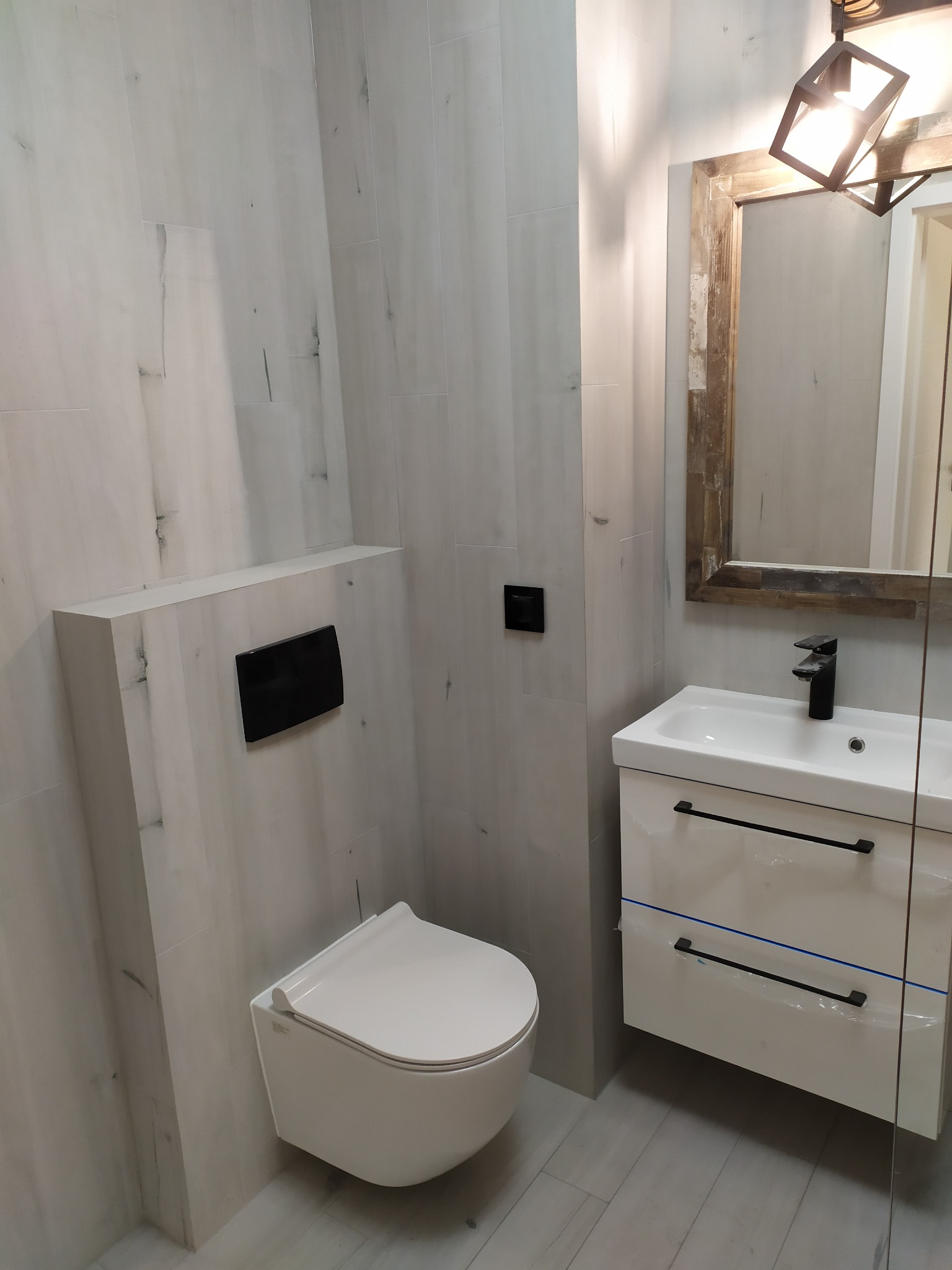 Nowoczesna łazienka z podwieszaną toaletą, umywalką z szafką w kolorze złamanej bieli i lustrem w drewnianej ramie, wykończona płytkami imitującymi drewno.