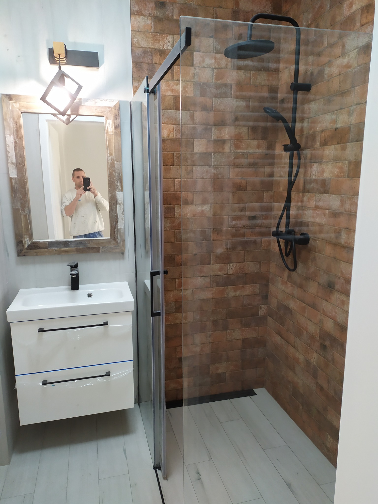 Wykończona łazienka w Warszawie z kabiną prysznicową typu walk-in z czarnymi elementami, umywalką z szafką w bieli oraz lustrem z drewnianą ramą, w której odbija się osoba robiąca zdjęcie.