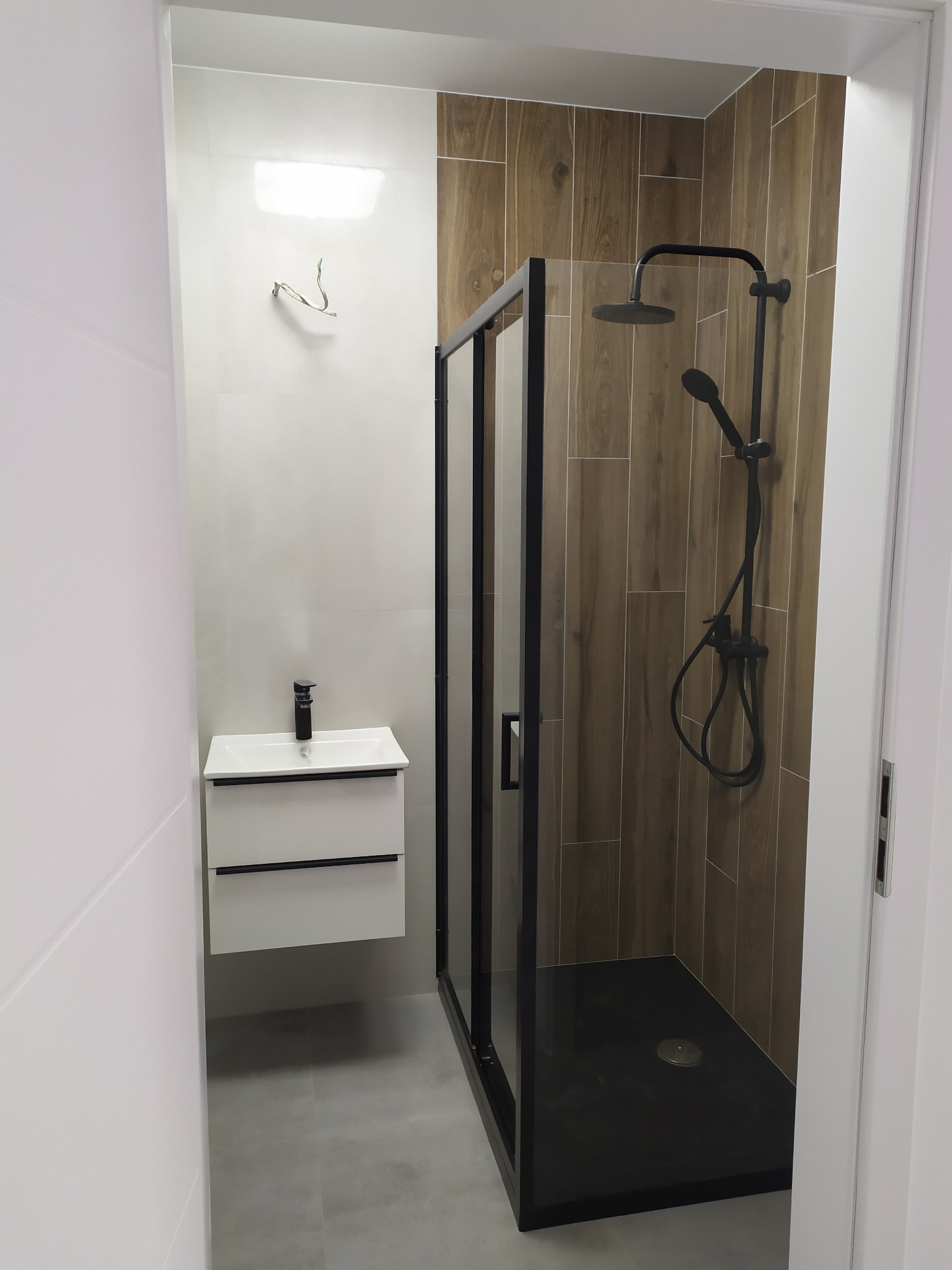 Nowoczesna łazienka z kabiną prysznicową typu walk-in z czarnymi profilami, umywalką z szafką w kolorze białym i drewnopodobnymi płytkami na ścianie w strefie prysznica.