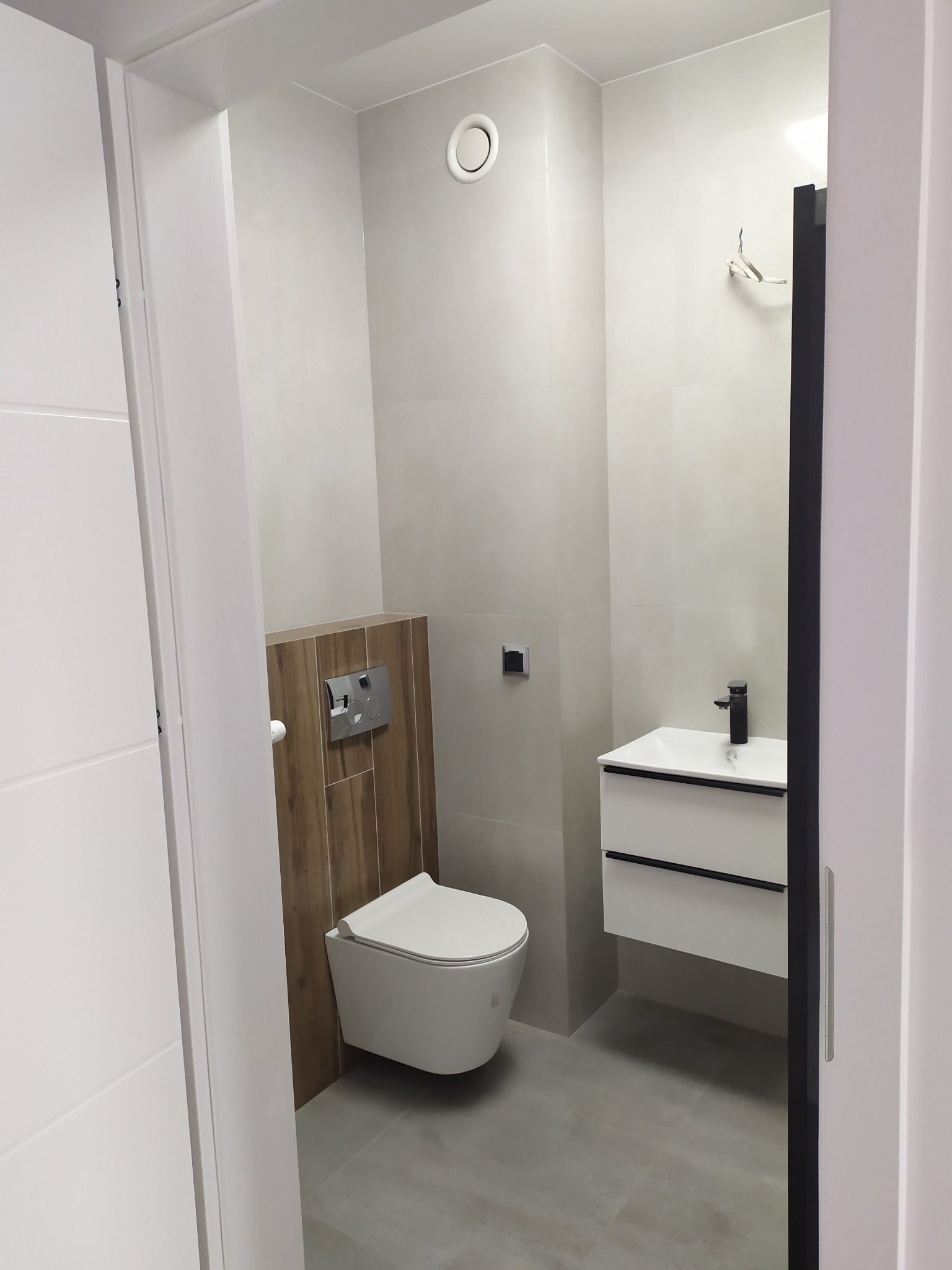 Nowoczesna łazienka z podwieszaną toaletą, umywalką z szafką i szarą płytką podłogową, widziana zza uchylonych białych drzwi.