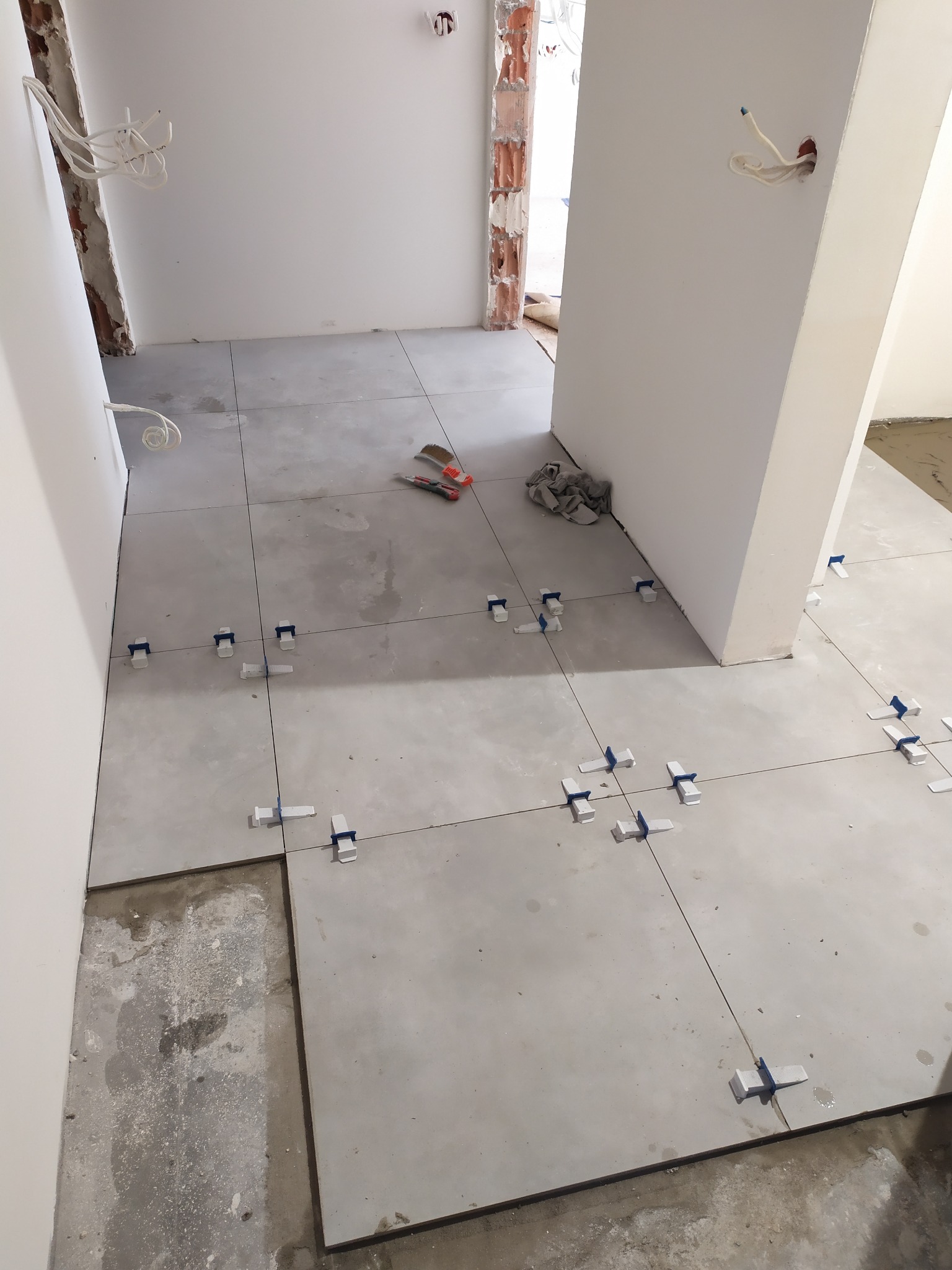 Układanie szarych płytek podłogowych z użyciem poziomujących klipsów w trakcie prac wykończeniowych, widoczne narzędzia i pozostałości zaprawy.