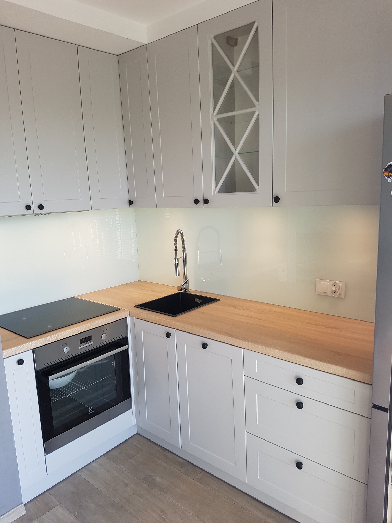 Jasnoszara zabudowa kuchenna z drewnianym blatem, czarnym zlewem i płytą indukcyjną, górne szafki z przeszklonymi frontami w geometryczne wzory.