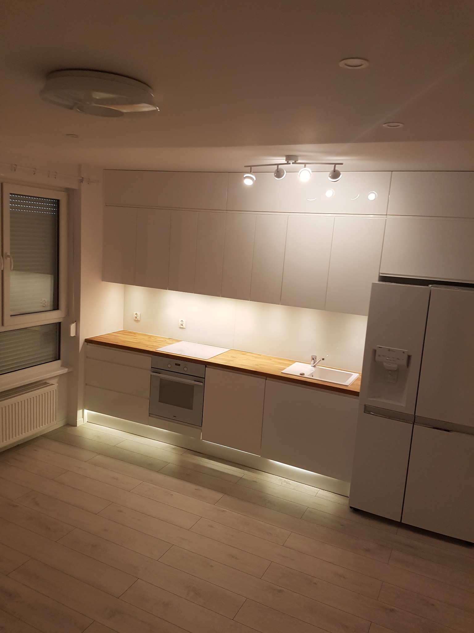 Nowoczesna kuchnia z białymi szafkami, drewnianym blatem i oświetleniem LED pod szafkami, widok na okno z żaluzjami i białą lodówkę.