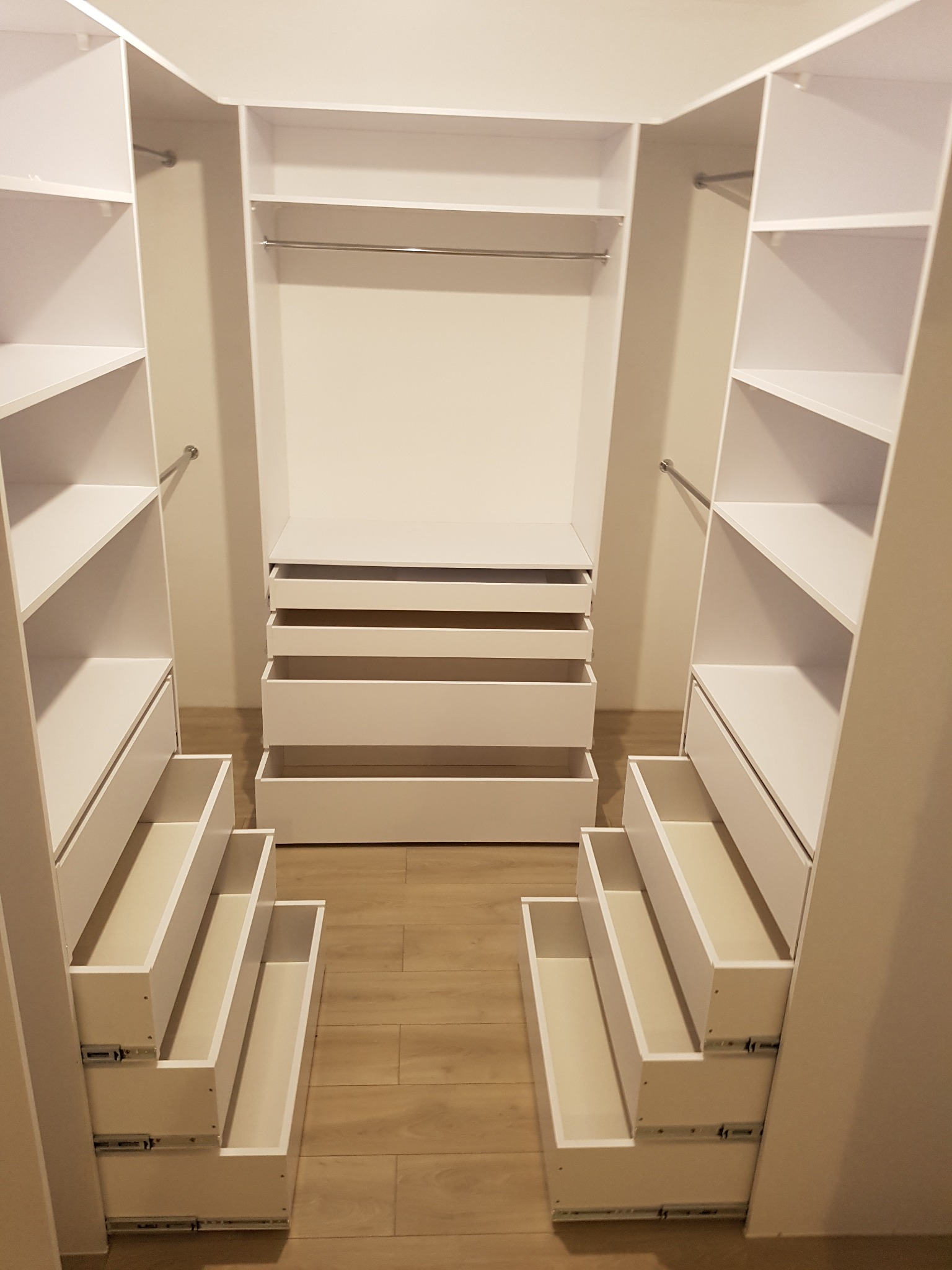 Wnętrze garderoby na wymiar z otwartymi szufladami, białe półki i drążki na ubrania, widok z perspektywy wejścia.