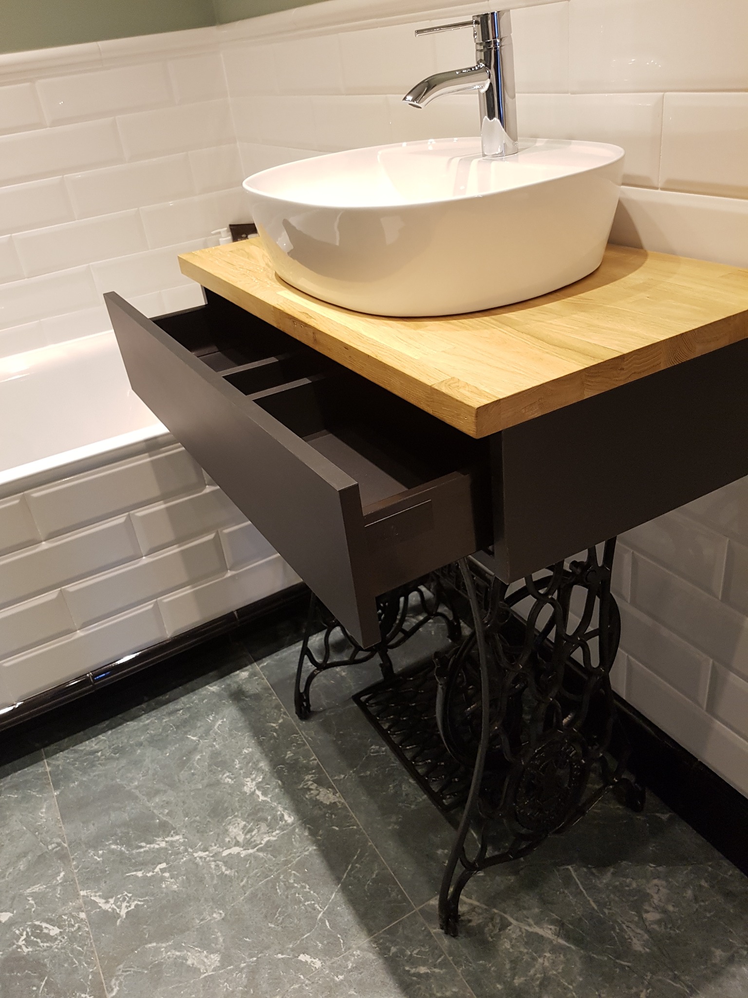 Umywalka nablatowa na blacie z jasnego drewna, zamontowana na metalowej podstawie starej maszyny do szycia, z wysuniętą szufladą, na tle białych płytek metra i marmurowej podłogi.