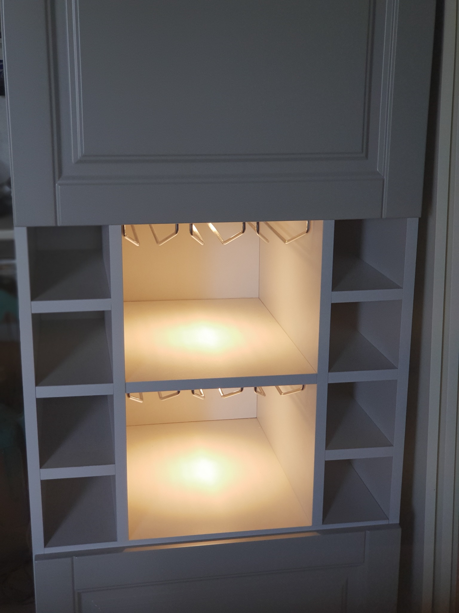 Szara szafka na wymiar z oświetlonymi półkami i uchwytami na kieliszki, widoczne boczne półki na wino.