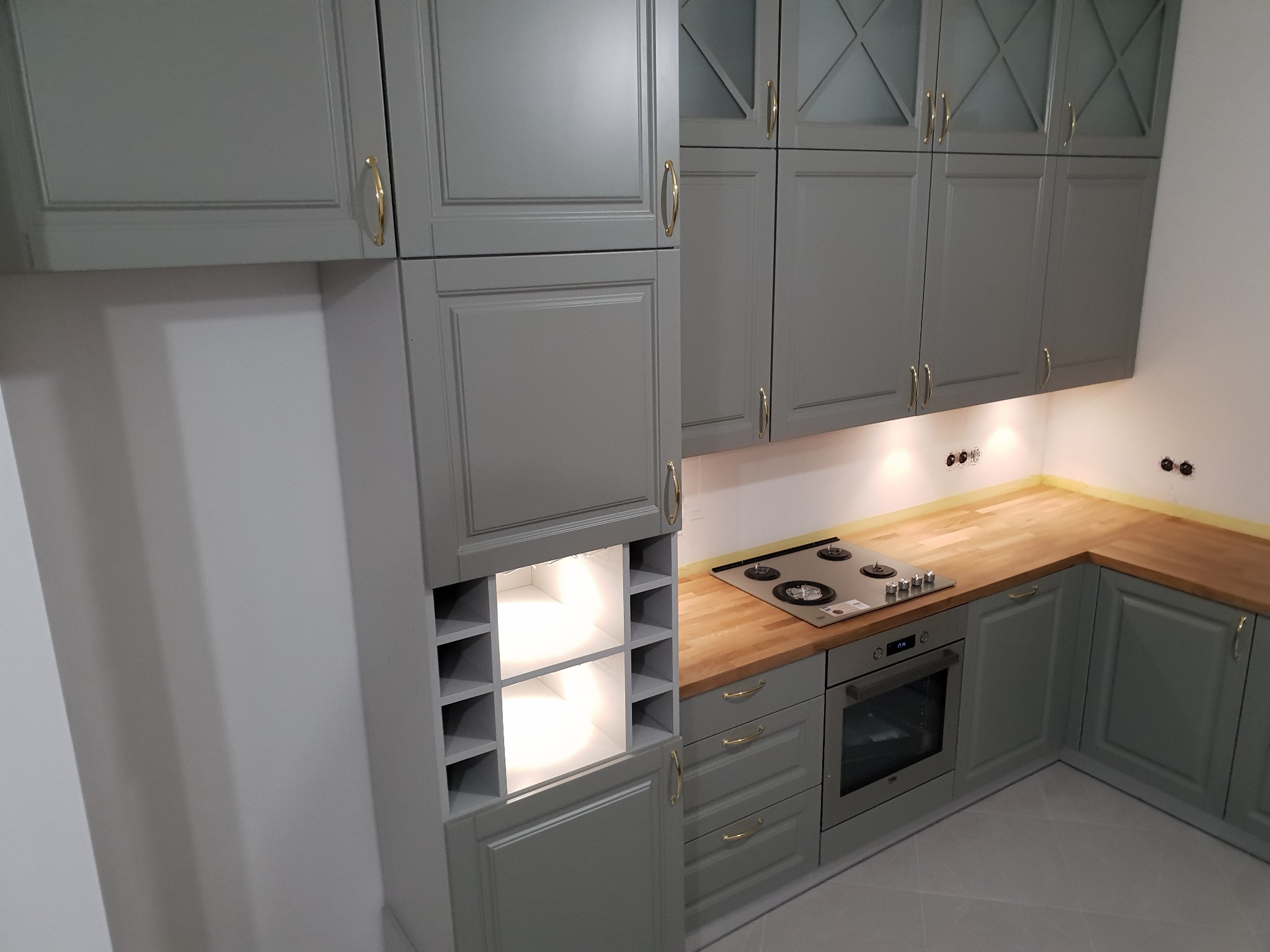 Szare szafki kuchenne z drewnianym blatem, złotymi uchwytami i wbudowaną półką na wino z podświetleniem LED.