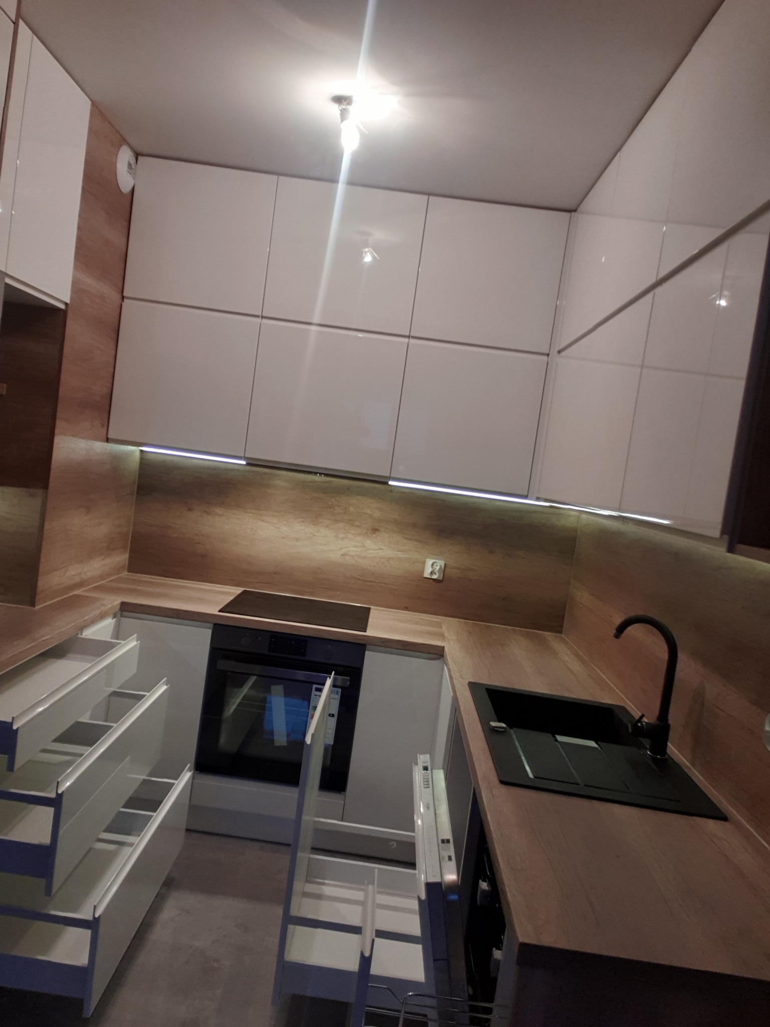 Nowoczesna kuchnia z białymi, lakierowanymi szafkami, drewnopodobnym blatem i podświetleniem LED pod szafkami wiszącymi, z wysuniętymi szufladami.