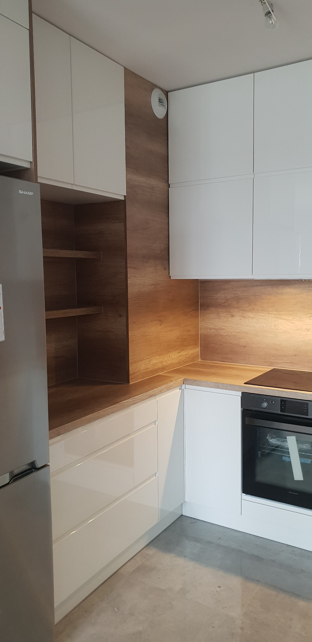 Biała, lakierowana zabudowa kuchenna z drewnianym akcentem na ścianie, widoczny fragment lodówki Sharp i piekarnika.