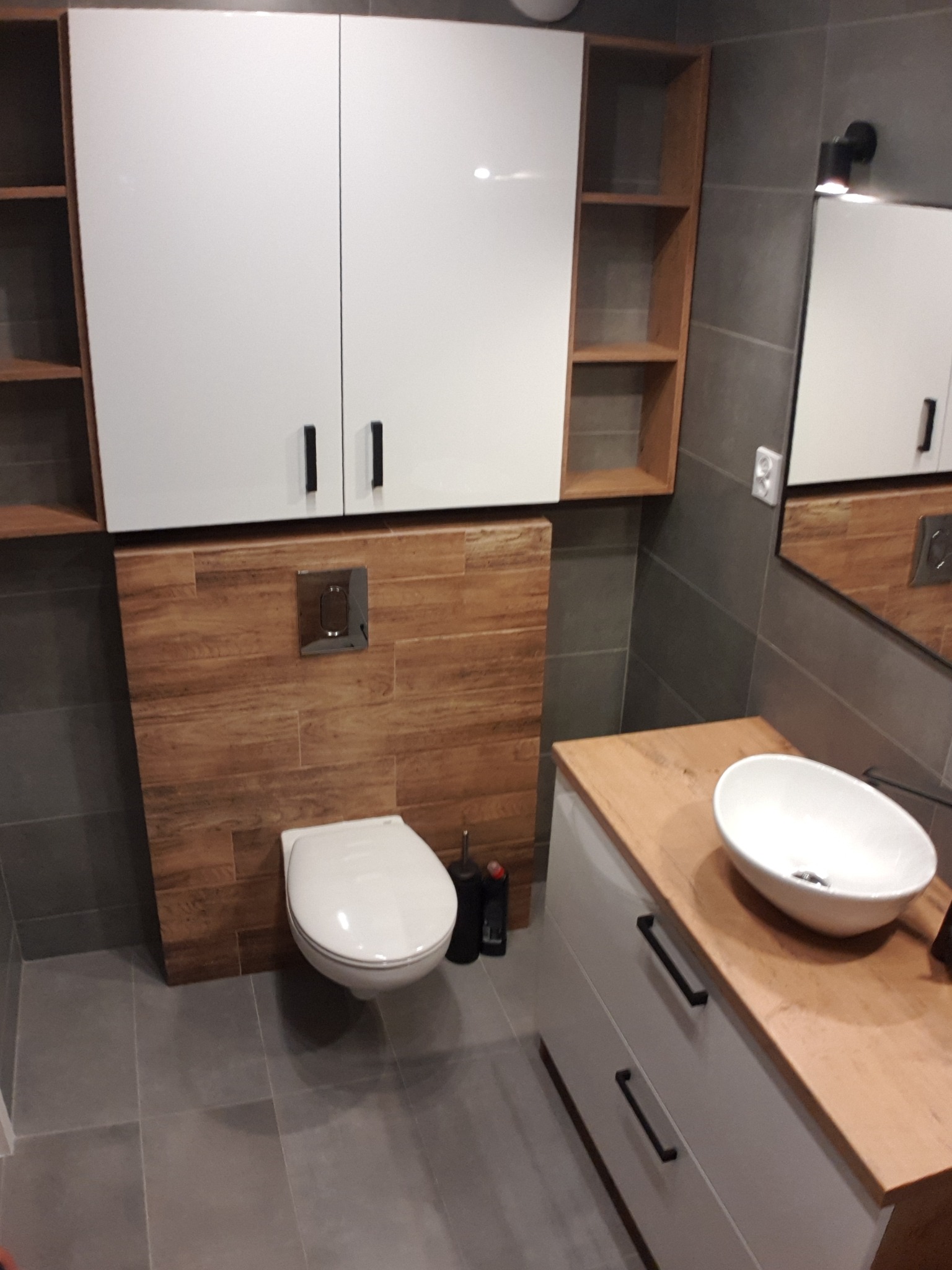 Nowoczesna łazienka z drewnianymi akcentami: szafki wiszące z białymi frontami, drewniane półki, umywalka nablatowa na szafce z drewnianym blatem, podwieszana toaleta z drewnianym panelem.