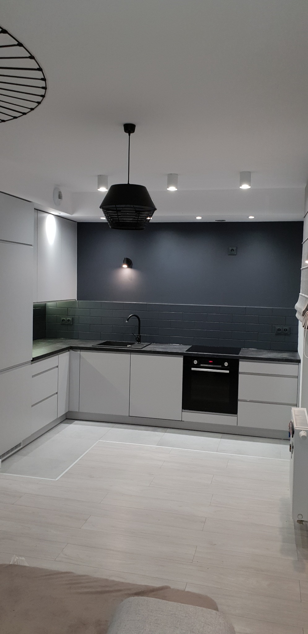 Nowoczesna kuchnia z białymi szafkami, czarnym zlewem i blatem, szarą ścianą z cegiełek oraz designerską lampą wiszącą z czarnego metalu.