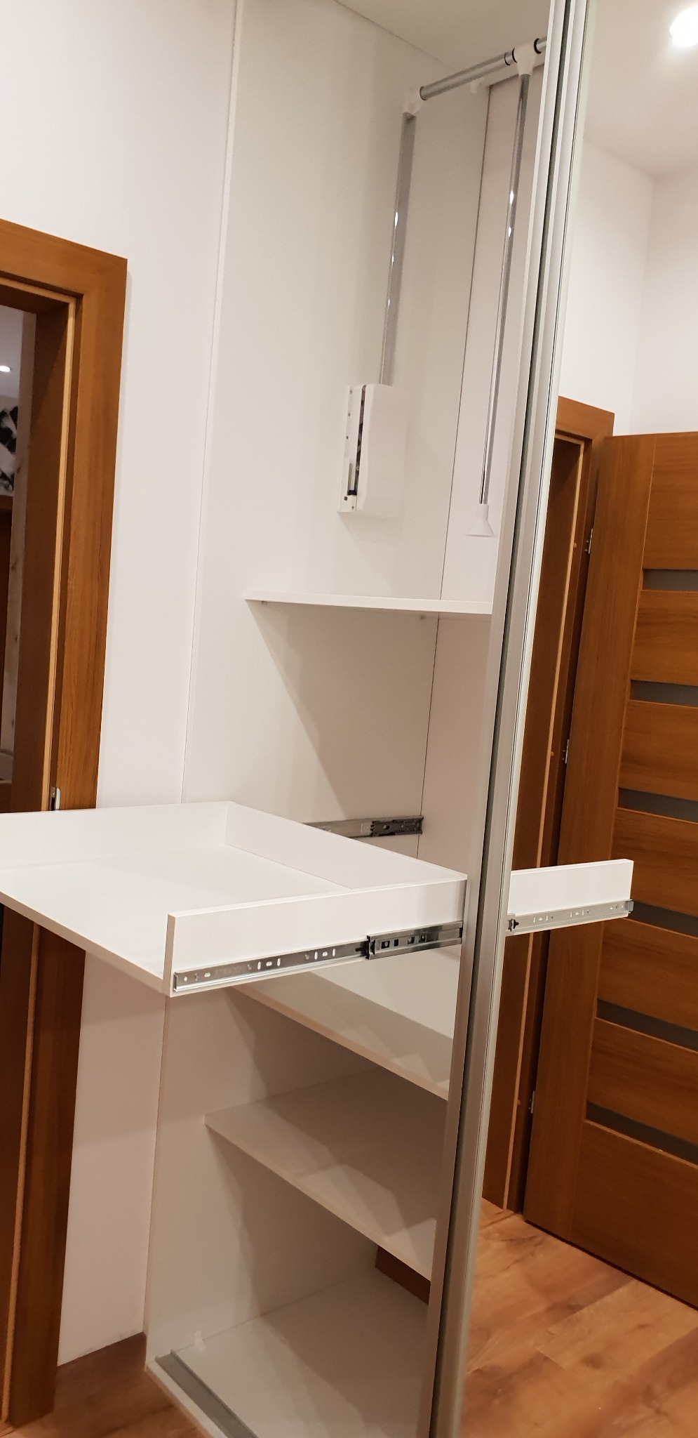 Wysuwana, biała półka w szafie na wymiar z widocznymi szynami, zamontowana w białej zabudowie, z elementami systemu przesuwnych drzwi i widoczną drewnianą ościeżnicą drzwi w tle.
