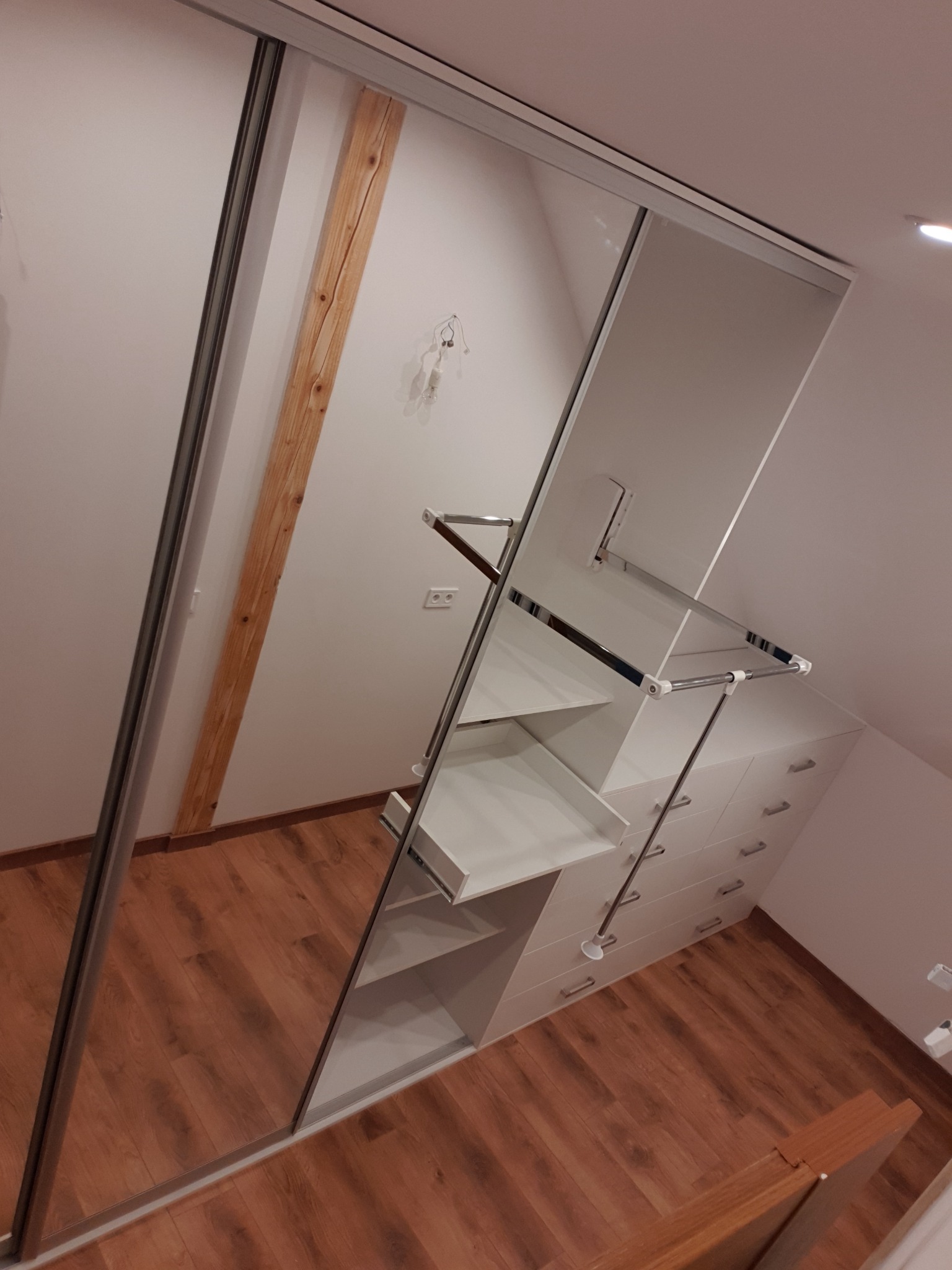 Biała szafa wnękowa z przesuwnymi lustrzanymi drzwiami i drewnianą belką stropową w tle, widoczne półki i szuflady.