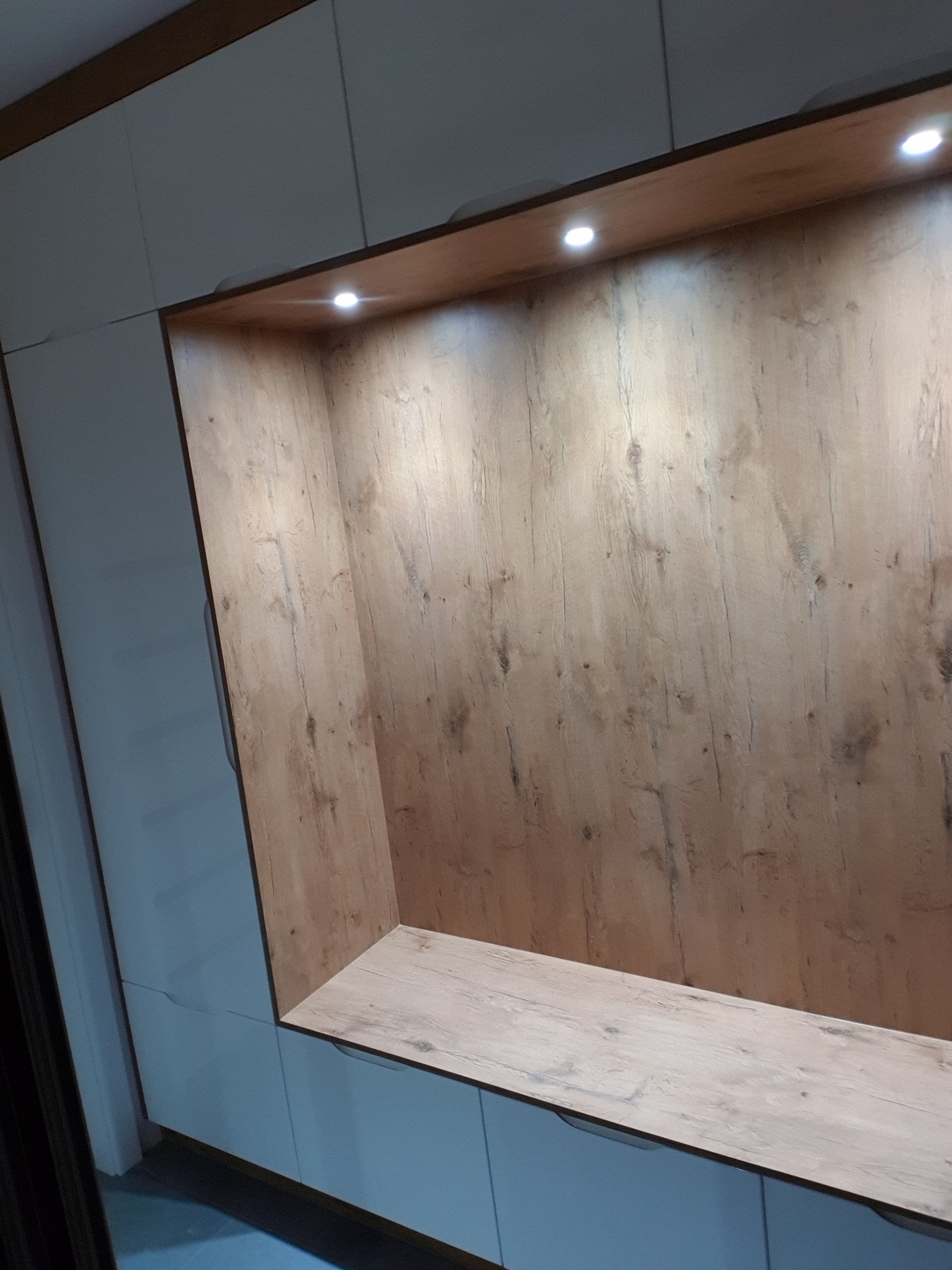 Wnęka w zabudowie meblowej z oświetleniem punktowym i wykończeniem imitującym drewno o jasnej barwie, z widoczną strukturą słojów.