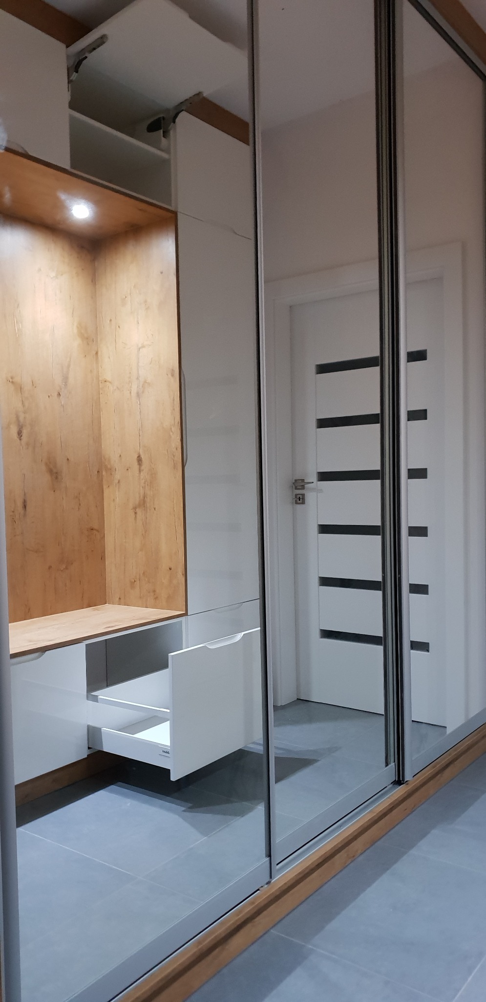 Wnętrze szafy wnękowej z lustrzanymi drzwiami przesuwnymi, widoczna otwarta szuflada z białym frontem i drewniane wykończenie wnęki z oświetleniem punktowym.