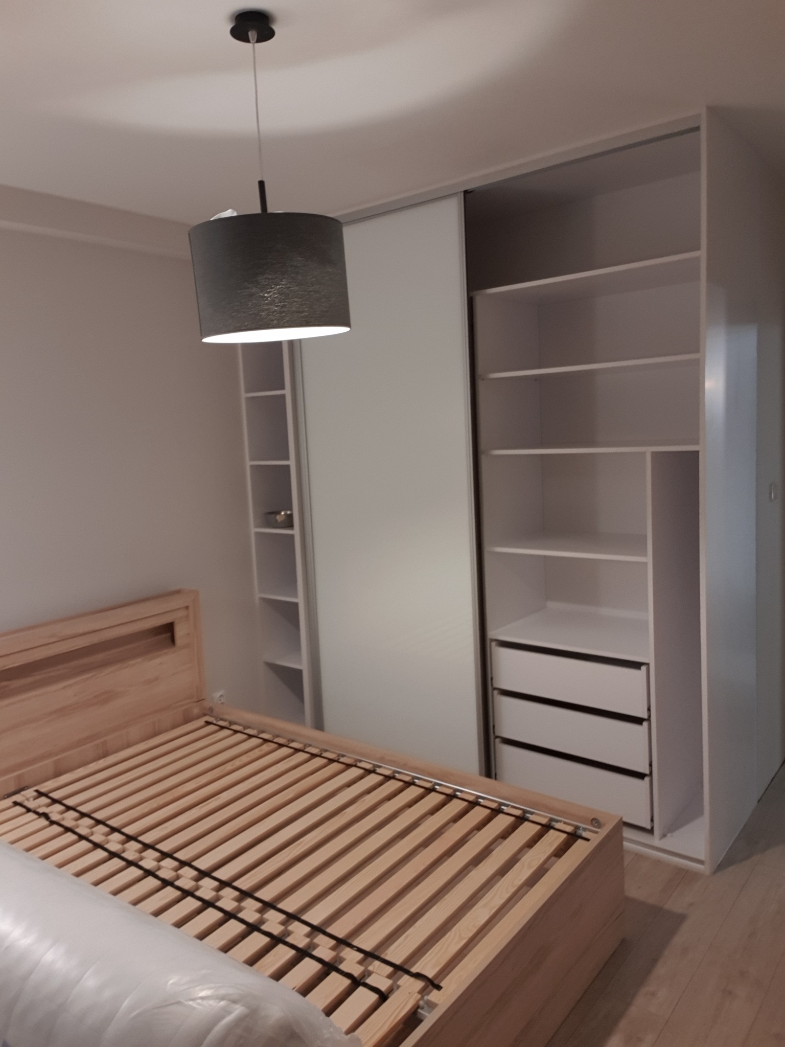 Szafa wnękowa z przesuwnymi drzwiami, jasne półki i szuflady, obok drewniane łóżko bez materaca, szary abażur lampy wiszącej.