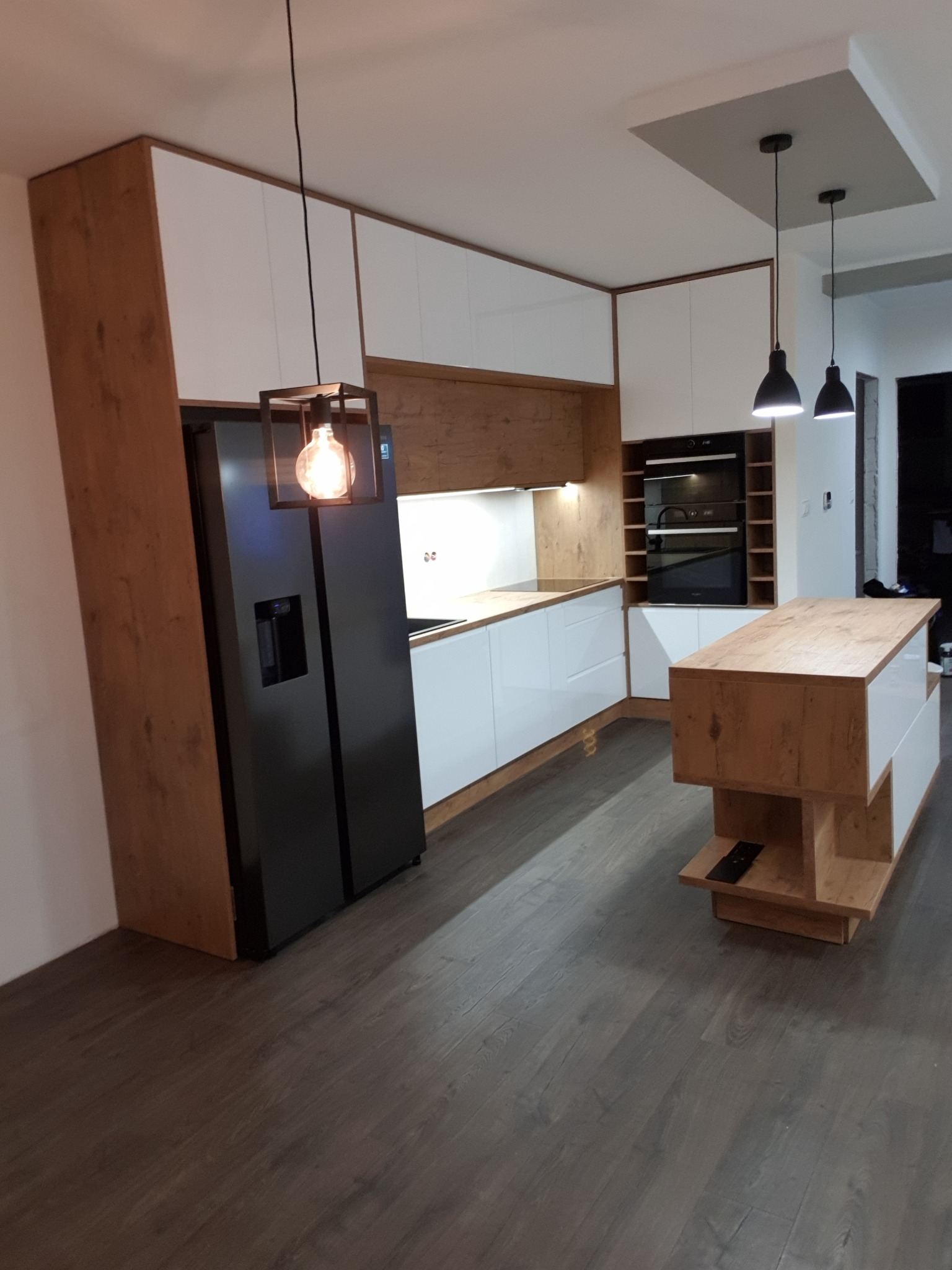 Nowoczesna kuchnia z białymi szafkami i drewnianymi akcentami, wyspą kuchenną oraz czarną lodówką side-by-side, oświetlona wiszącymi lampami z żarówkami Edisona.