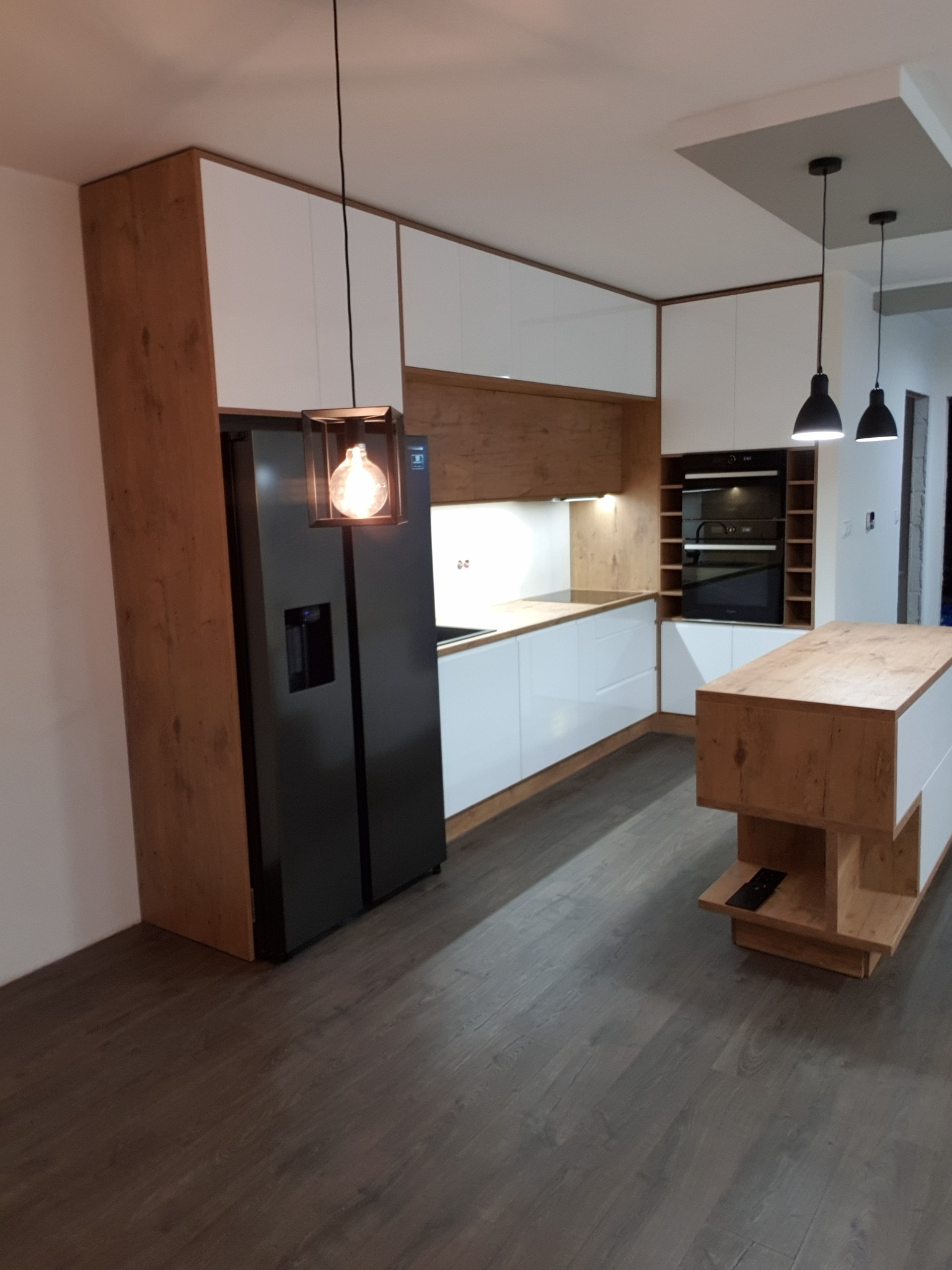 Nowoczesna kuchnia z białymi frontami i drewnianymi akcentami, w tym obudowa lodówki i wyspa kuchenna, oświetlona wiszącą lampą z żarówką Edisona.