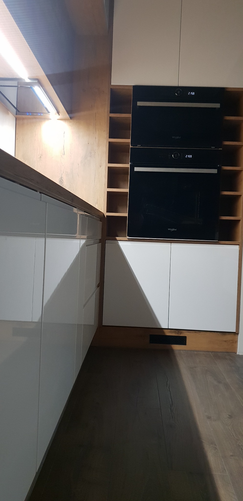 Nowoczesna kuchnia z białymi, lakierowanymi frontami, drewnianymi akcentami i wbudowanymi piekarnikami Whirlpool.